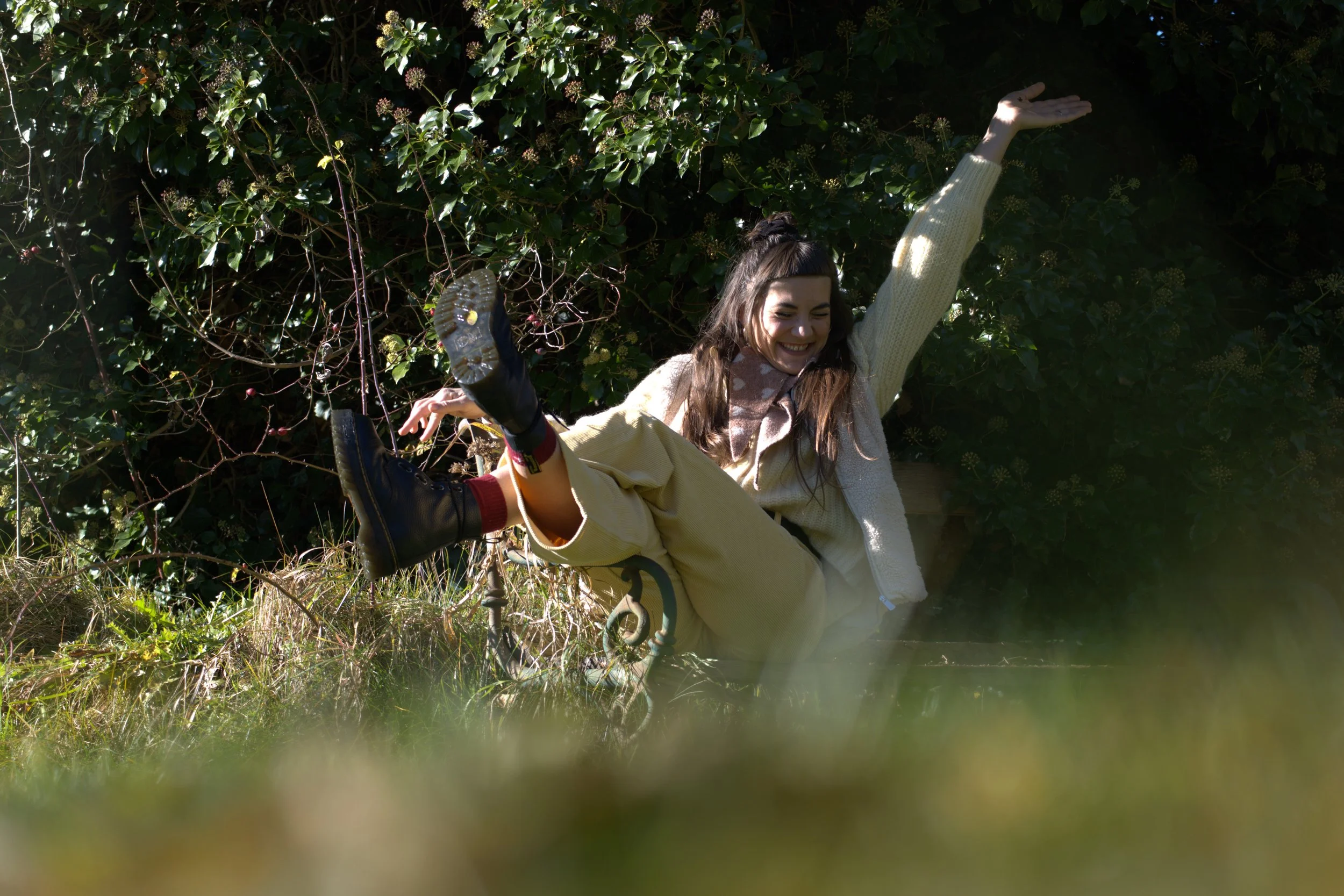 Junge Frau lacht lachend auf Rasen, mit einem Bein in der Luft, in einer grünen Gartenumgebung, sonniger Tag