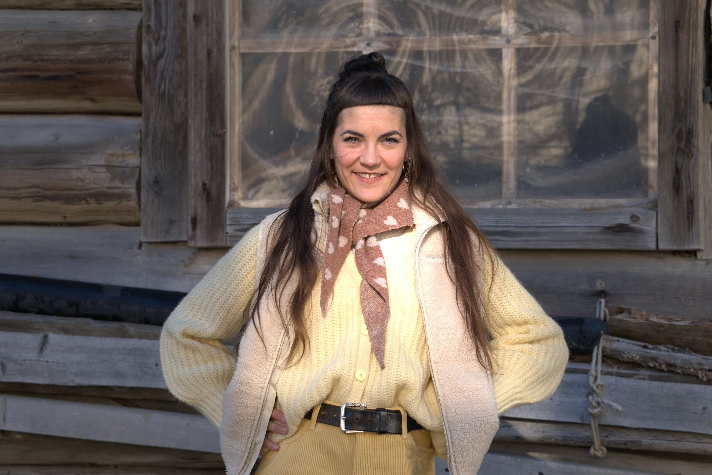 Frau lächelt vor einem Holzschuppen, trägt eine gelbe Jacke und einen braunen Schal mit weißen Herzen.