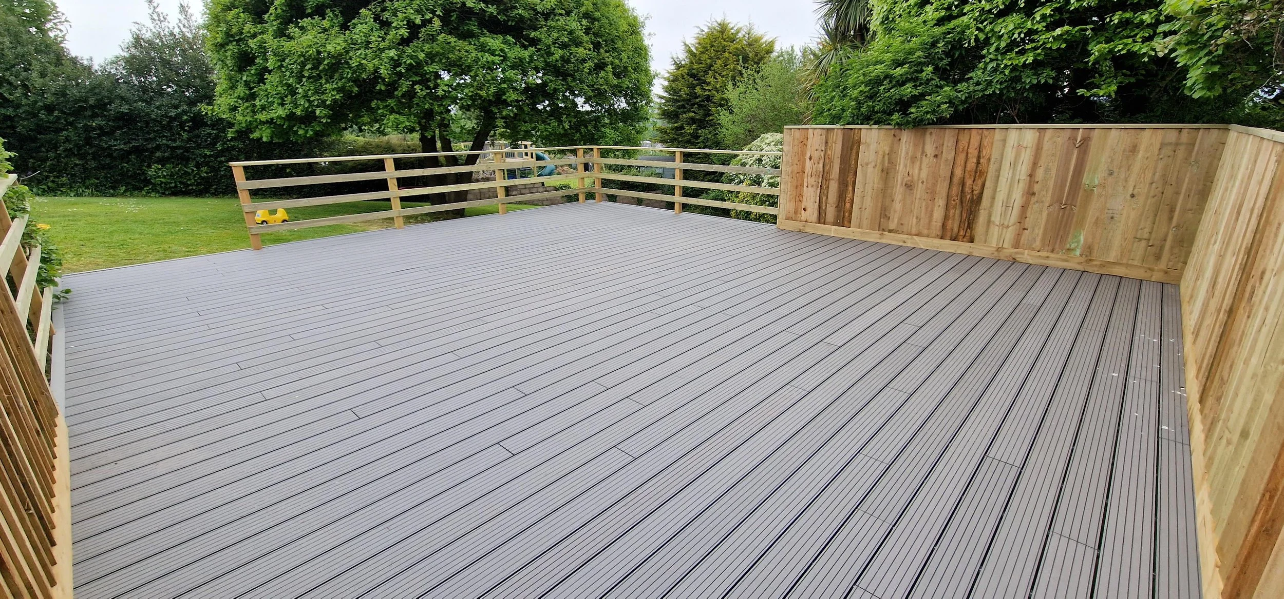 A newly built wooden deck with gray composite decking boards, enclosed by a wooden fence, in a backyard with green trees and grass.