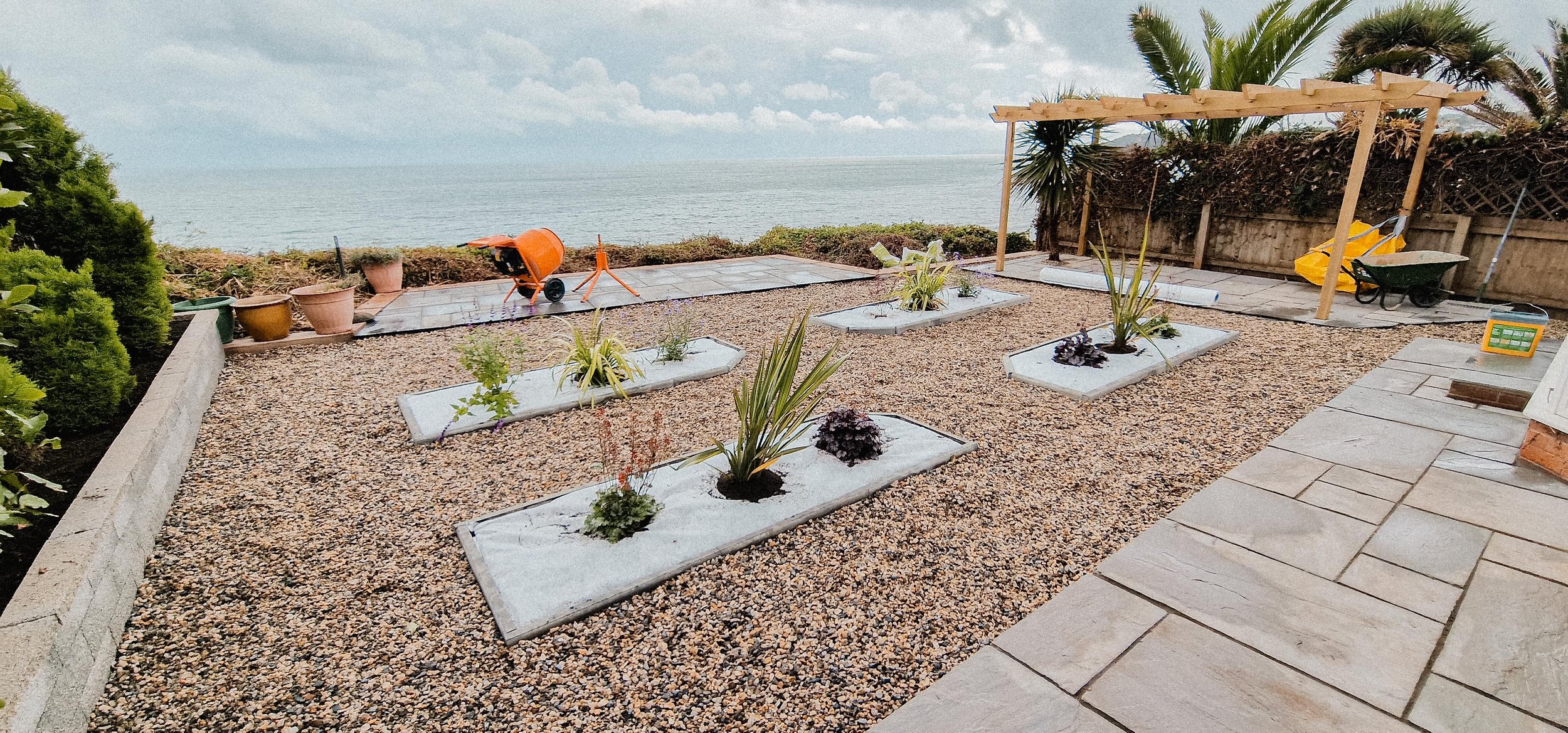 A landscaped backyard with raised garden beds containing plants, gravel ground cover, and a stone patio on the right. In the background, there is a view of the ocean with a cloudy sky. Construction materials and tools, including an orange cement mixe