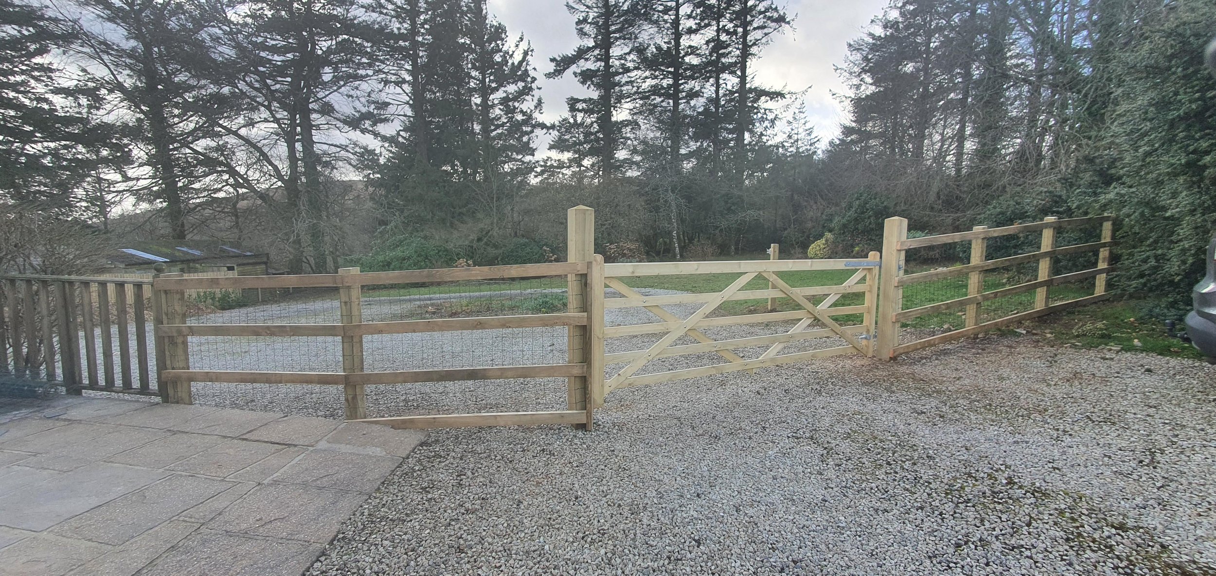 Wooden gate and fence in a yard with gravel ground, trees and bushes in the background.