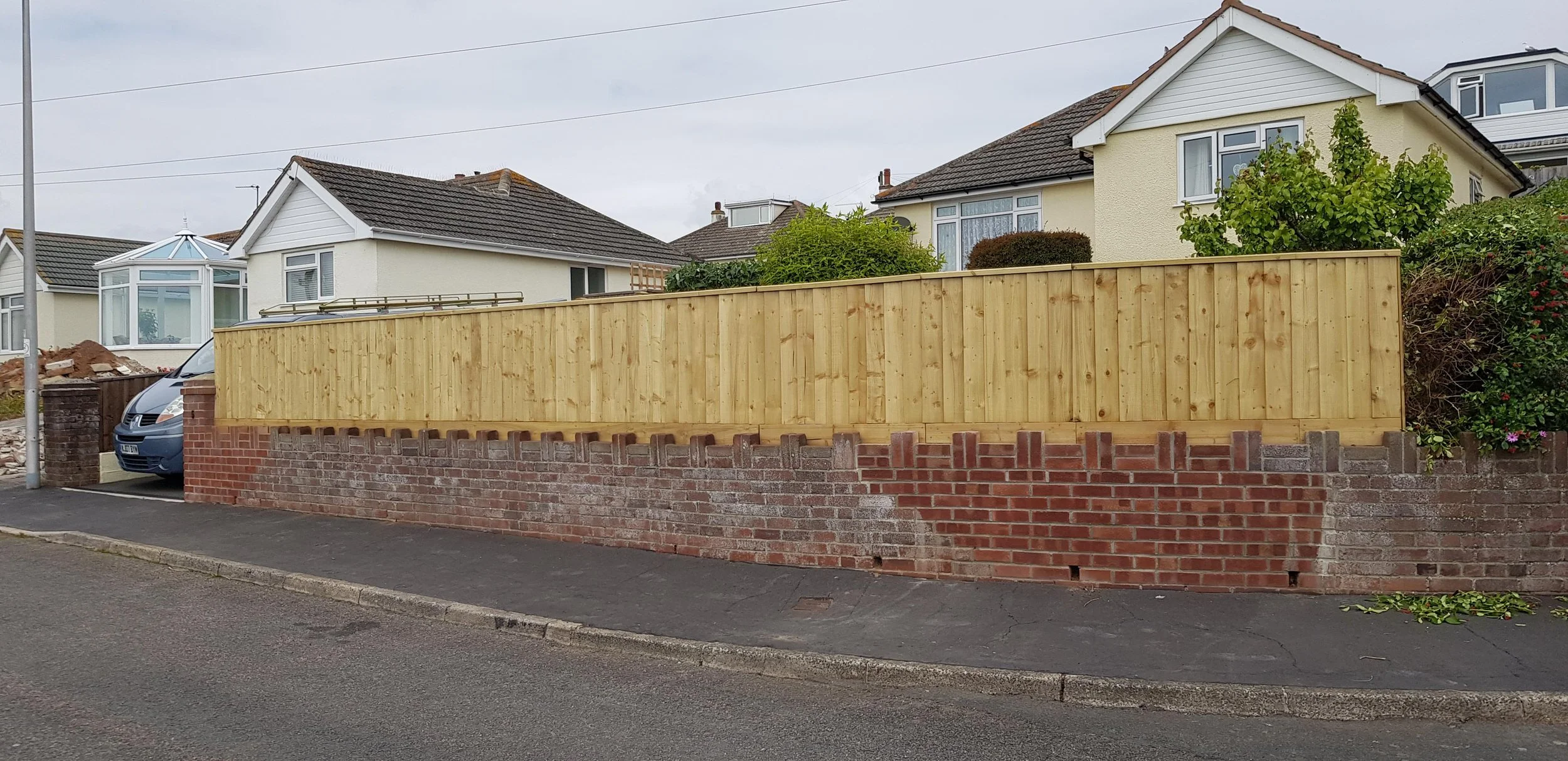 Residential street with a house partially hidden by a brick and wooden fence. The house has a tiled roof and a bay window, with some bushes visible behind the fence.