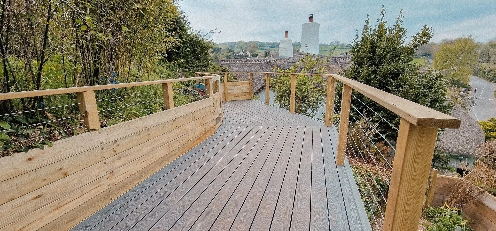 A wooden outdoor deck with a railing, located on a hillside surrounded by trees, overlooking buildings and a distant landscape with chimneys and rolling hills.