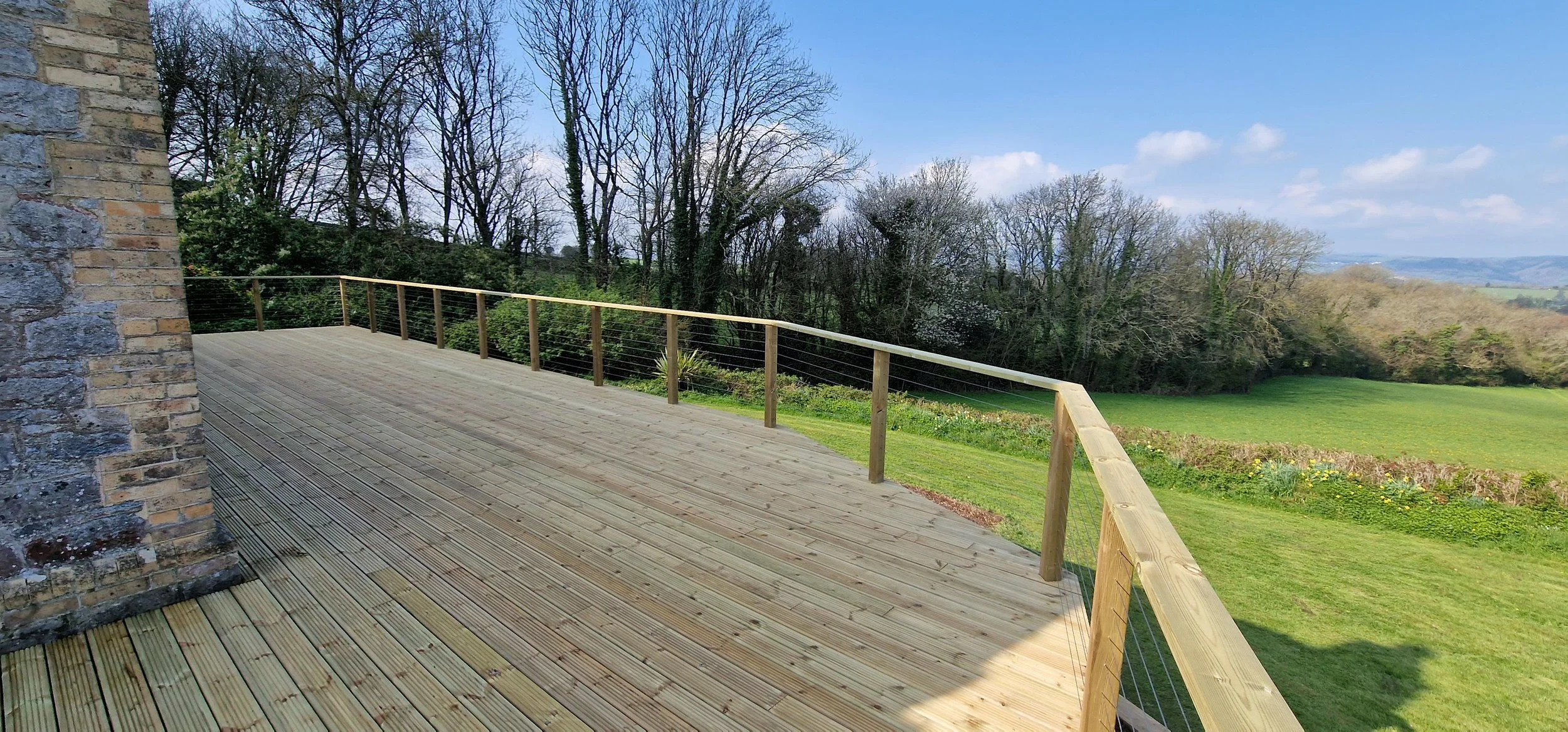 A wooden terrace with a rope and wood railing overlooking a grassy field and trees in the distance on a clear, sunny day.