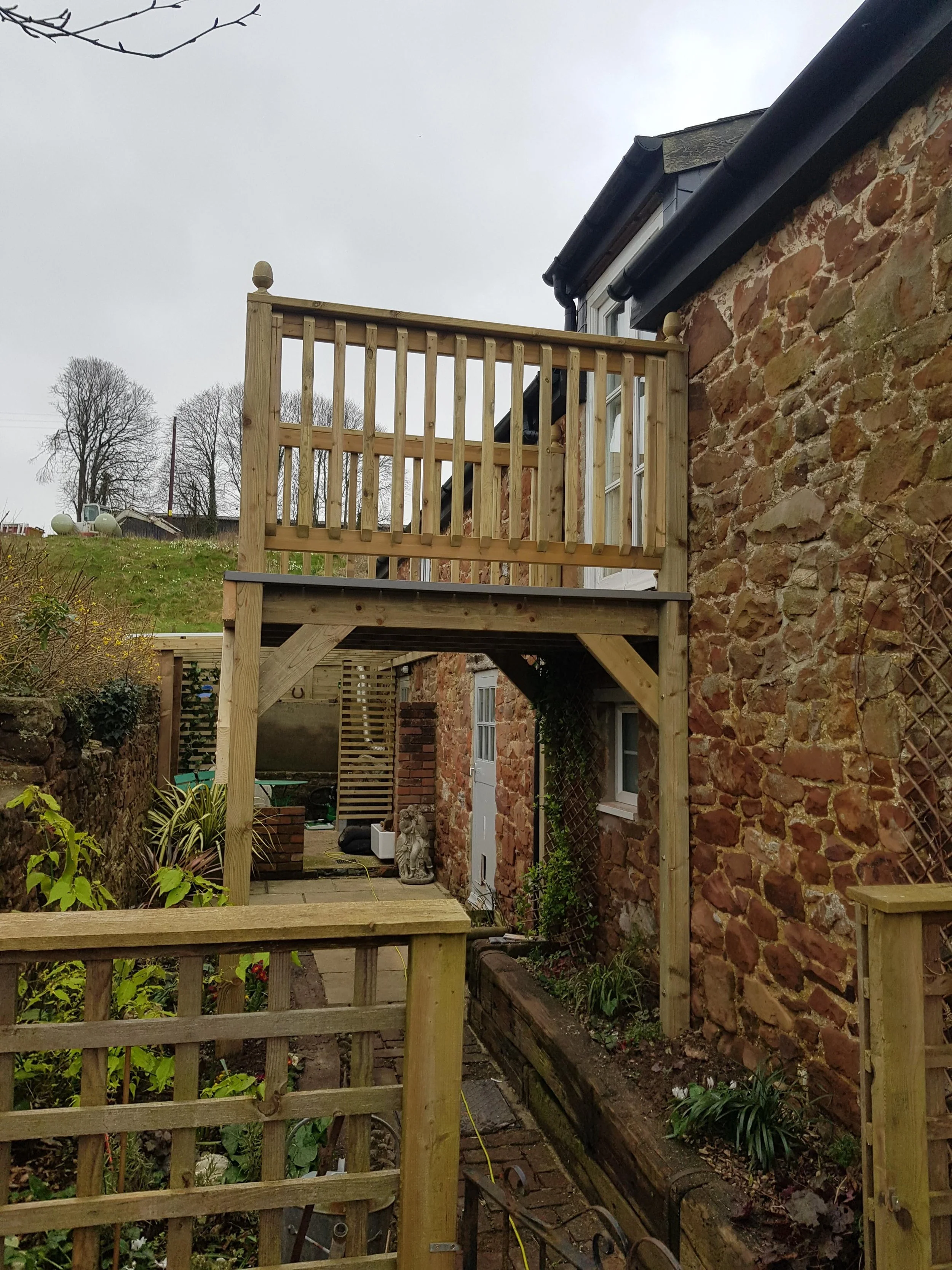 Wooden balcony with railings attached to a brick house, overlooking a garden area with plants and paving stones.