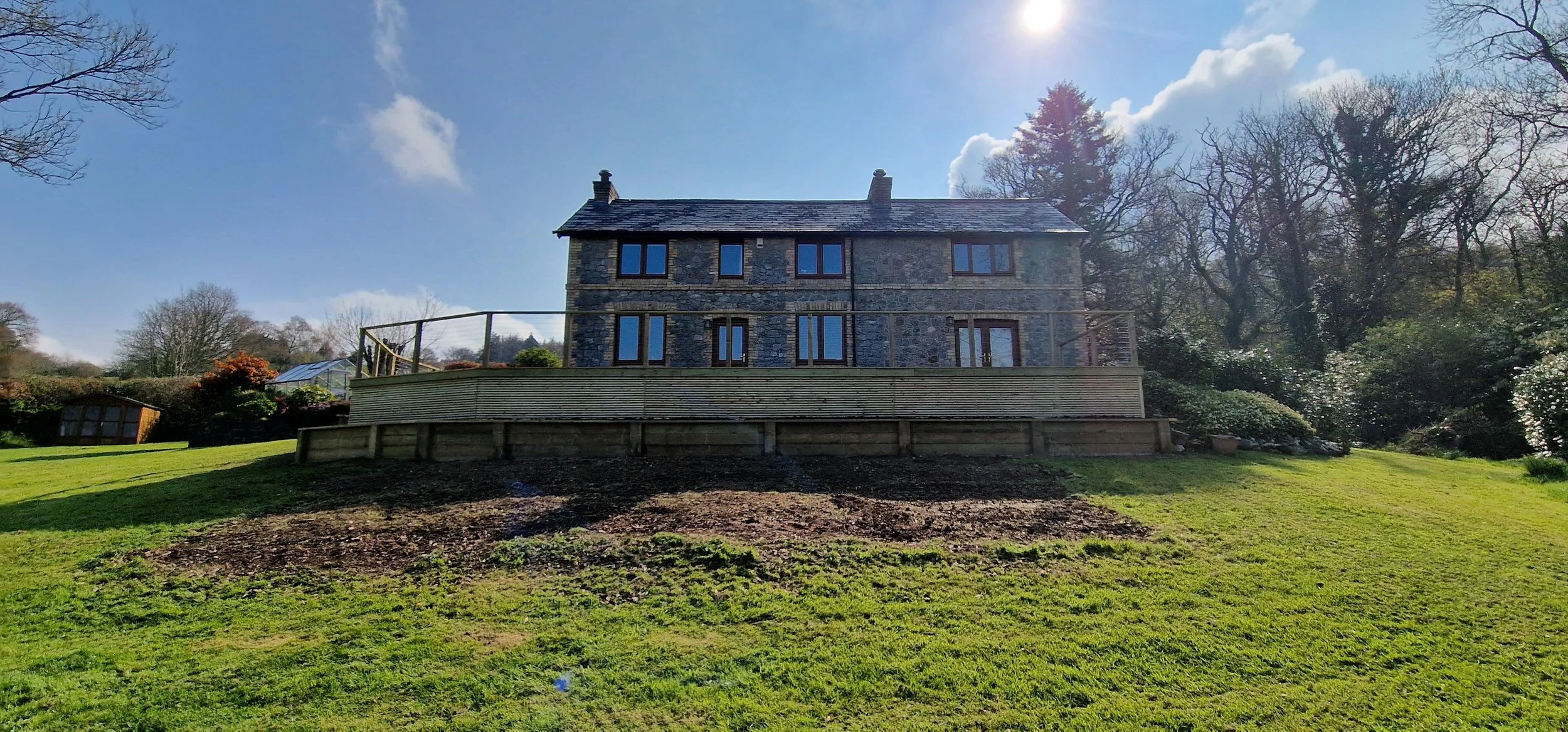 a stone house on a grassy hill with trees in the background, partly cloudy sky, and sunlight shining through the clouds