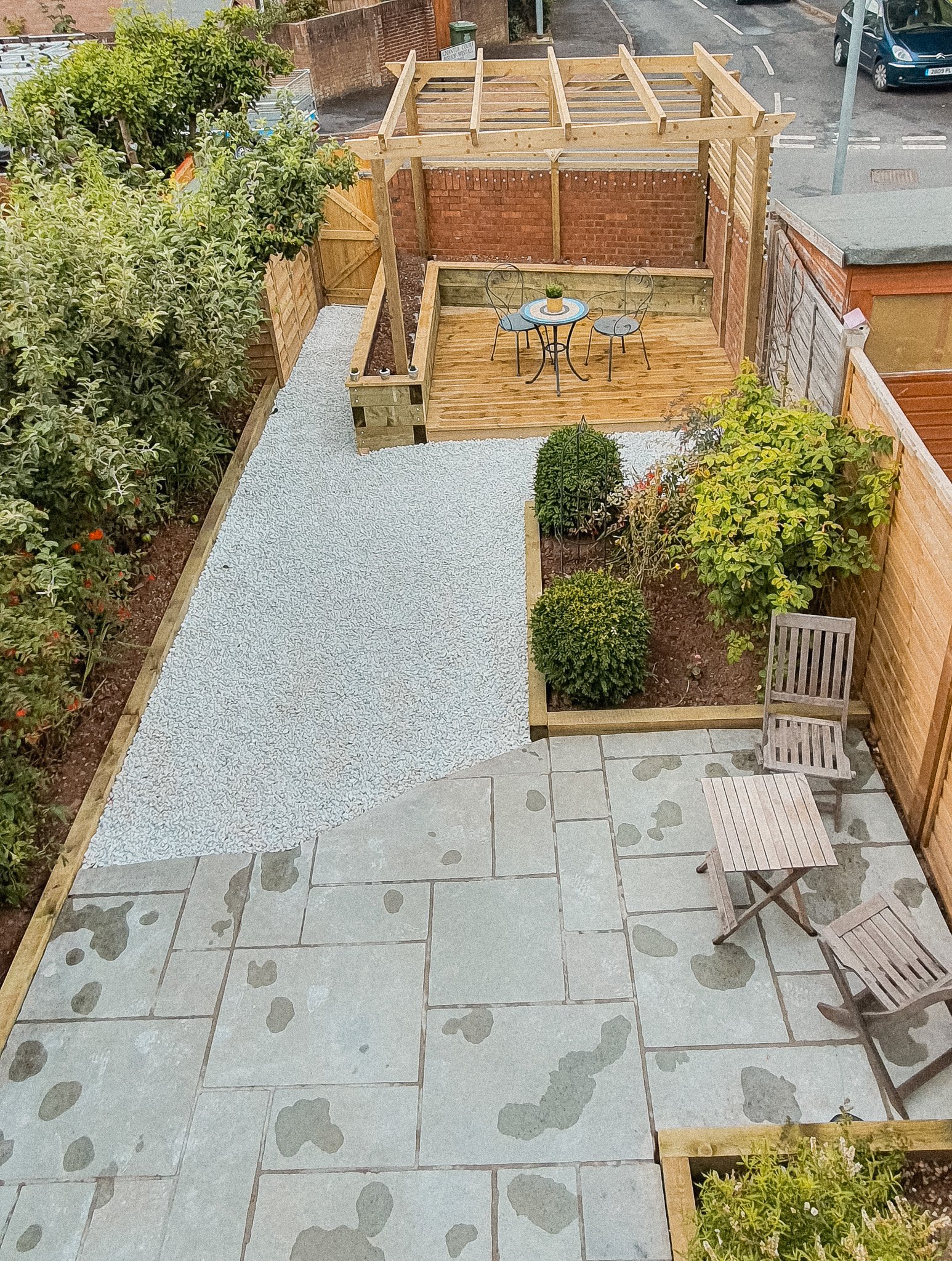 A backyard with a gravel path, a tiled patio with chairs and a small table, a wooden deck with a table and two chairs, and a garden area with plants and trees, all bordered by wooden fences.