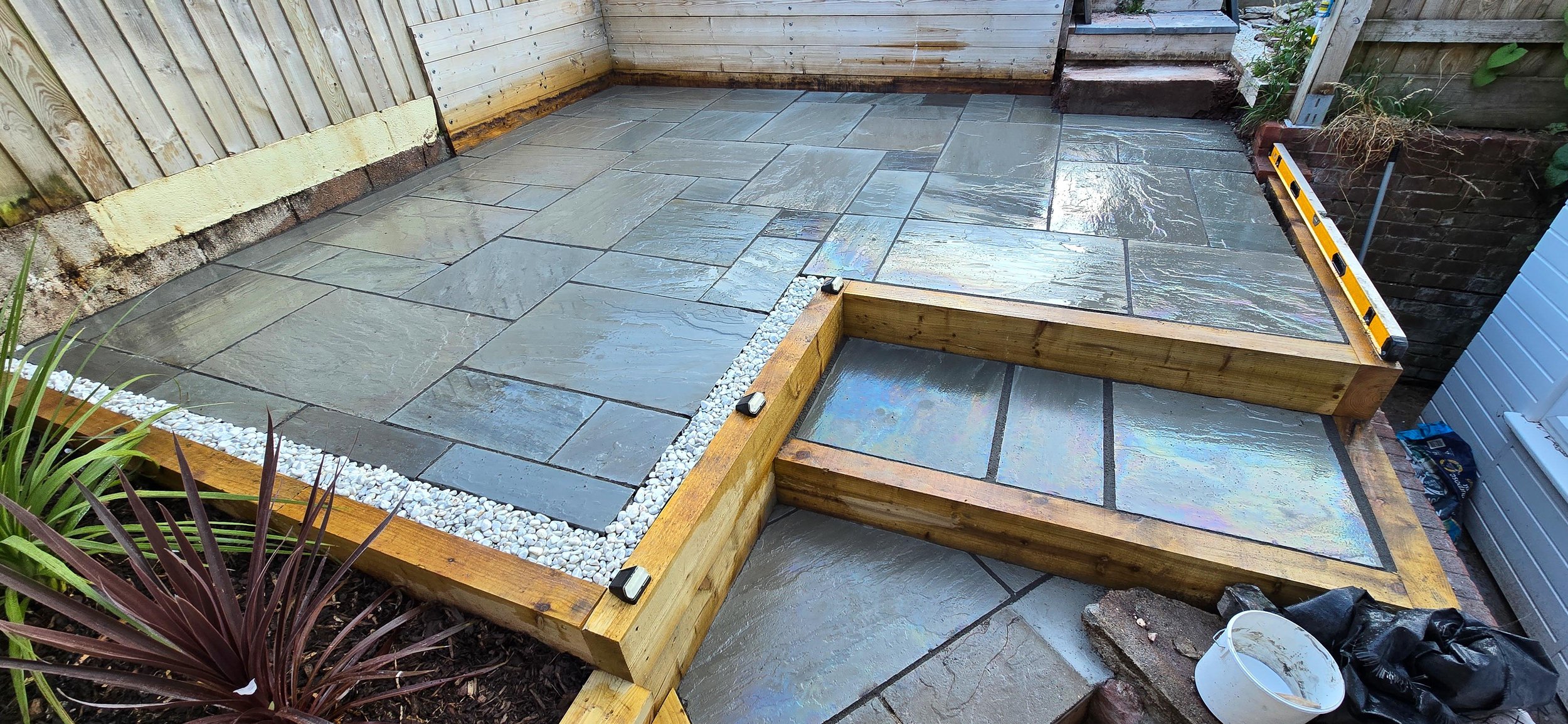 A partially completed outdoor patio with stone tiles, some of which are still being installed, surrounded by wooden framing and construction tools.