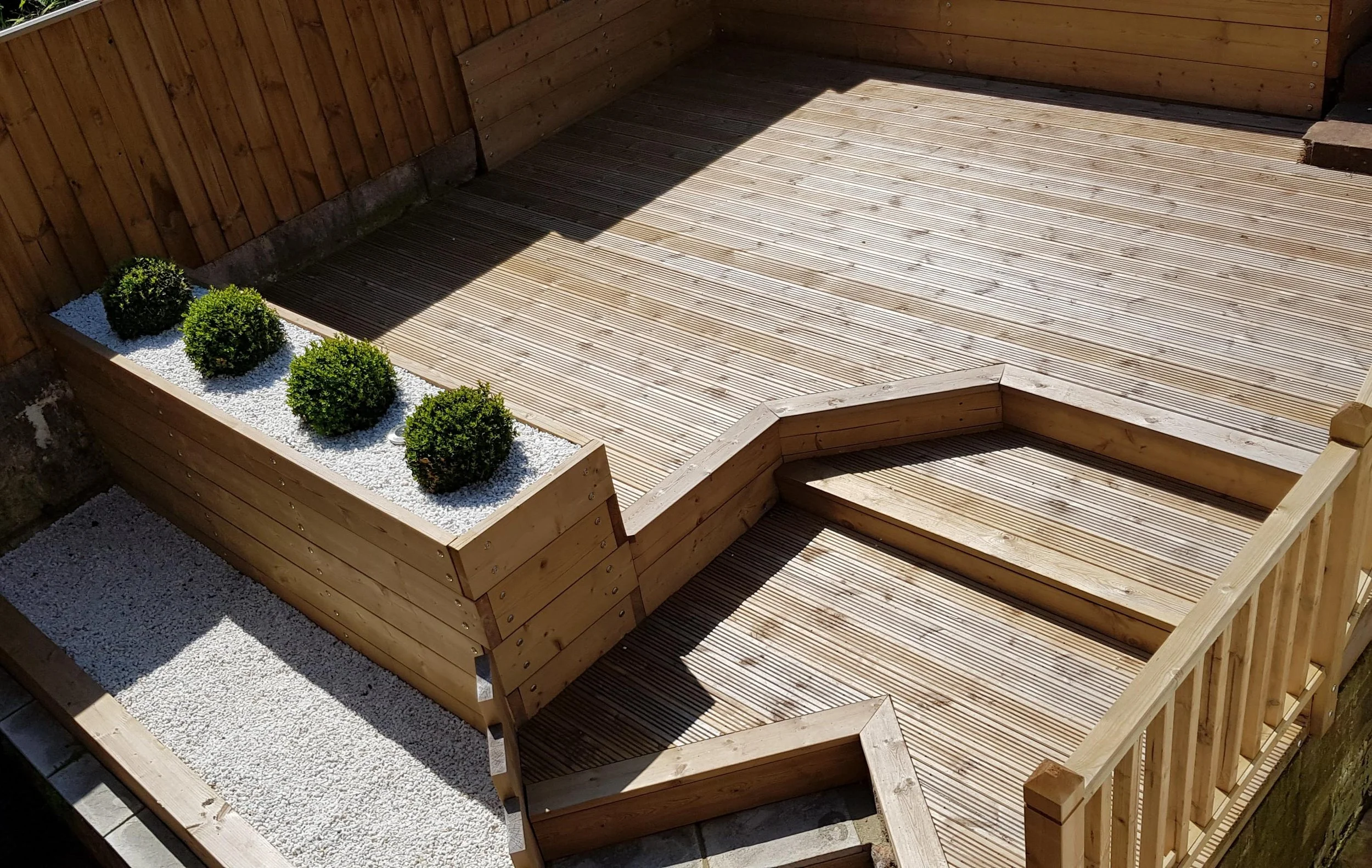 A wooden deck with stairs and a planter box containing four small green bushes, surrounded by white gravel.