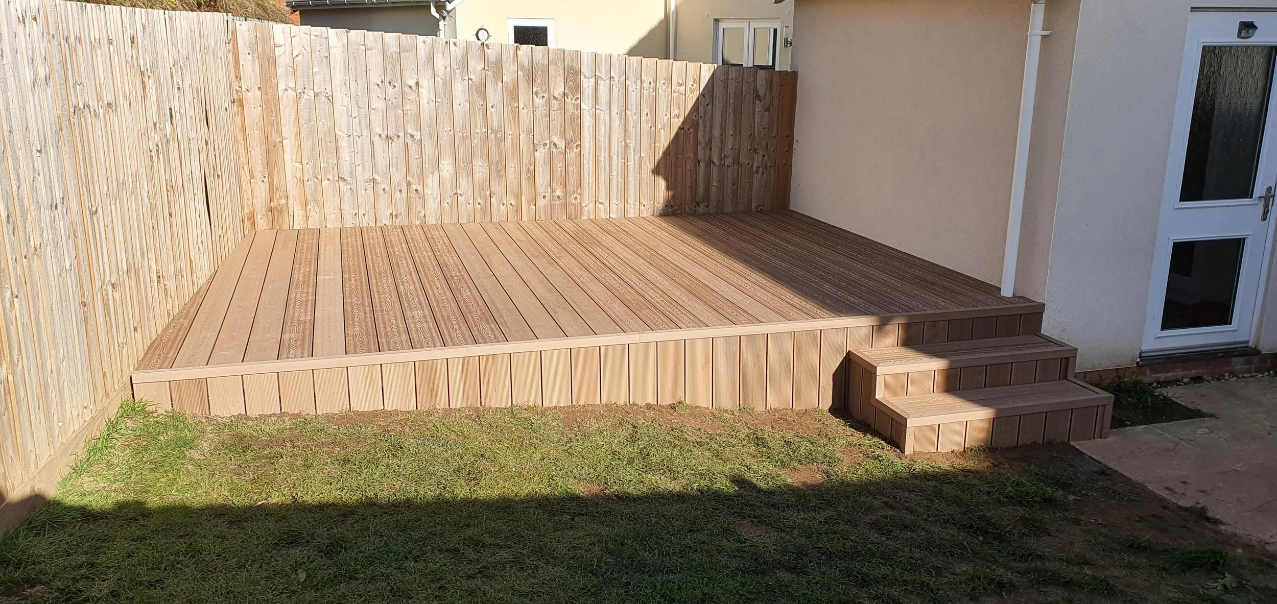 A newly built wooden deck with steps in a backyard, enclosed by a wooden fence, next to a house with a glass door.