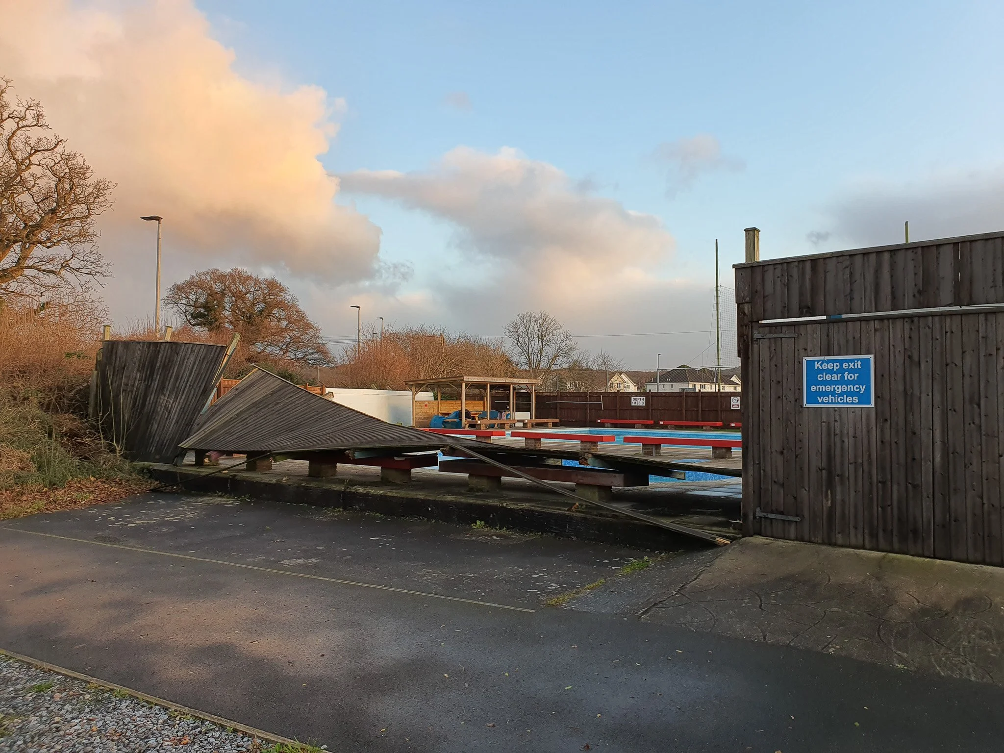A damaged wooden barrier and a fallen fence at a boat slip or dock area, with a sign to keep exit clear for emergency vehicles, and a car parking area in the foreground. The sky is partly cloudy.