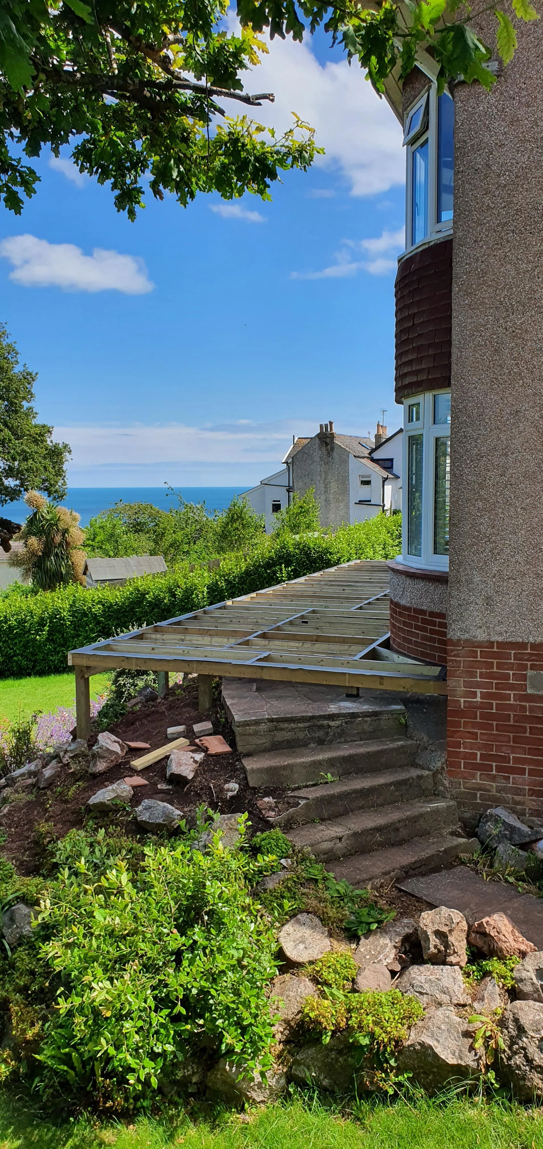 Side view of a house under construction with a new wooden deck frame, stone stairs leading up, surrounded by greenery and garden rocks, with neighboring houses and the ocean in the background under a partly cloudy sky.