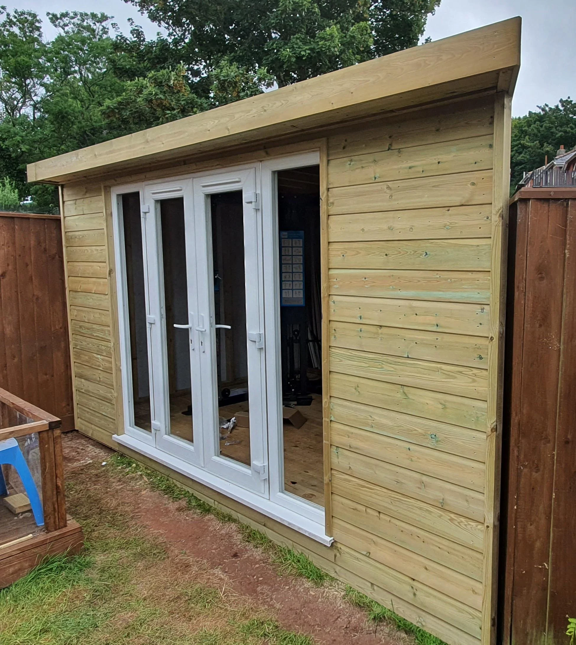 A wooden backyard shed with white double glass doors in a fenced yard, with trees in the background and a small garden bed with a blue chair on the left.