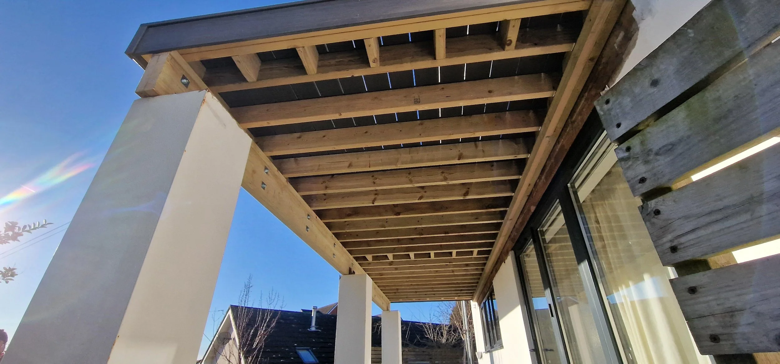 Under construction or renovation of a balcony or patio area with exposed wooden beams and support columns, next to a window with a curtain, in a residential neighborhood with rooftops and a clear blue sky.
