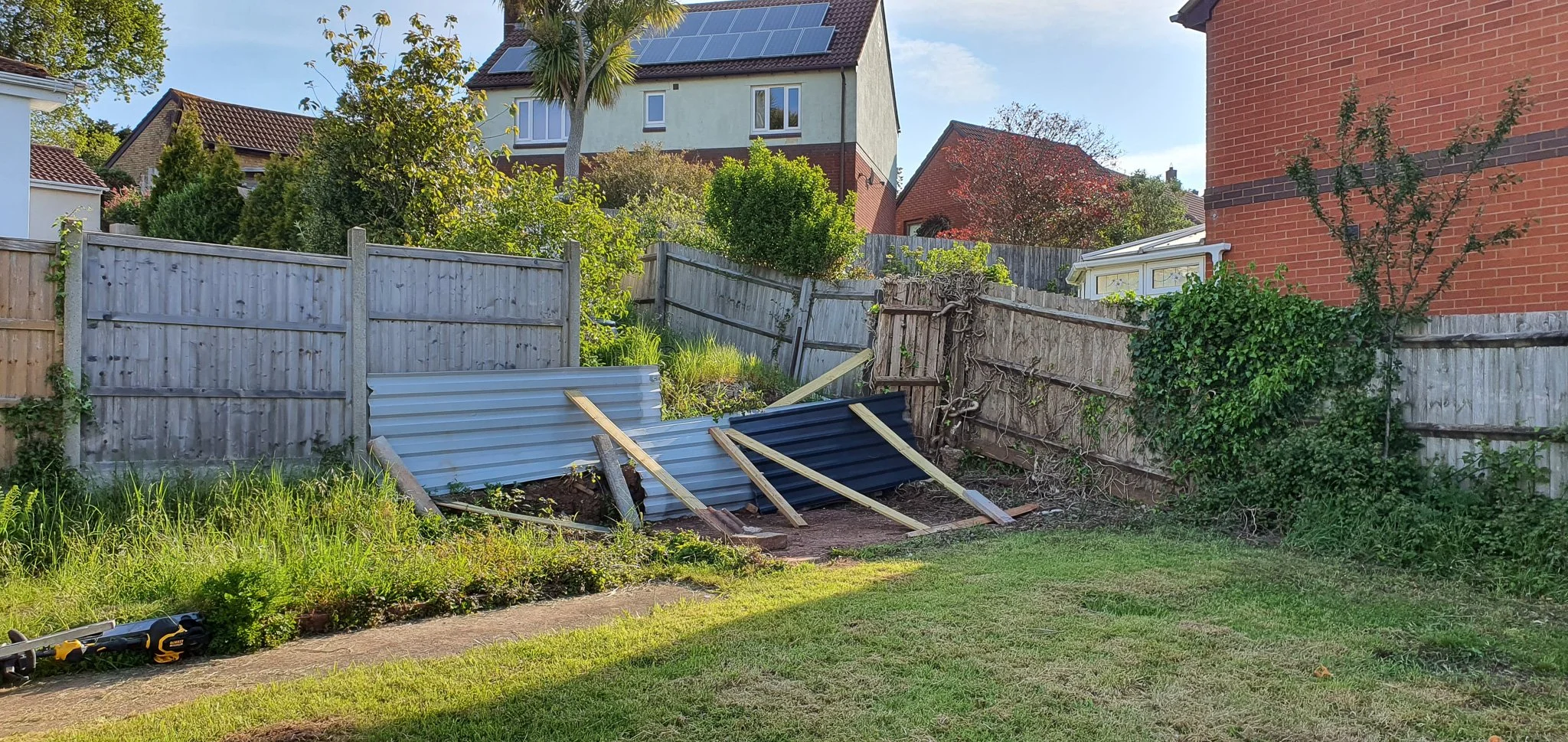 A backyard with a wooden and metal fence, some damaged corrugated metal sheets leaning against the fence, and construction tools on the ground. There are trees and bushes, and a neighboring house with solar panels on the roof in the background.