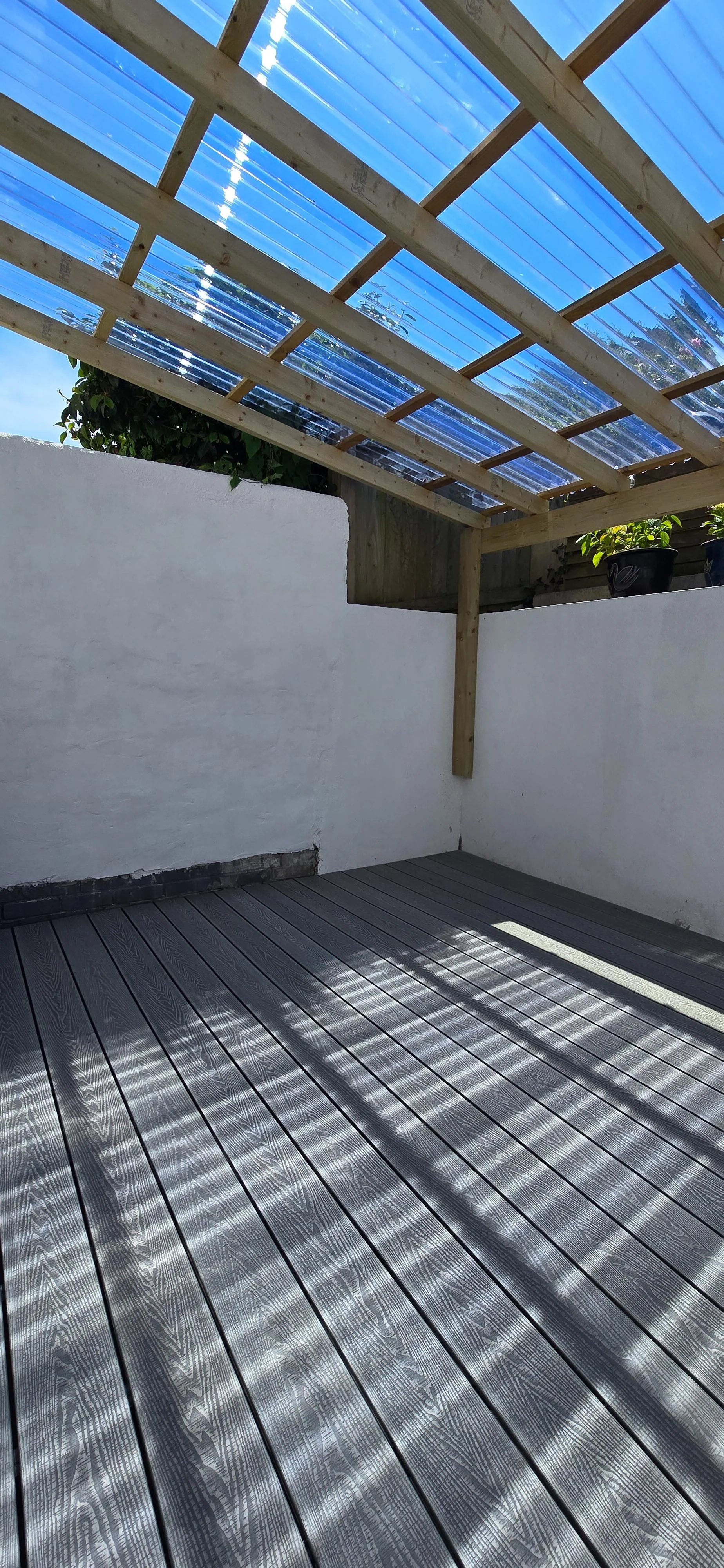 Wooden deck with shadow patterns, white exterior wall, partially constructed wooden and plastic roof framework with clear plastic panels, potted plants on a ledge beyond the wall.