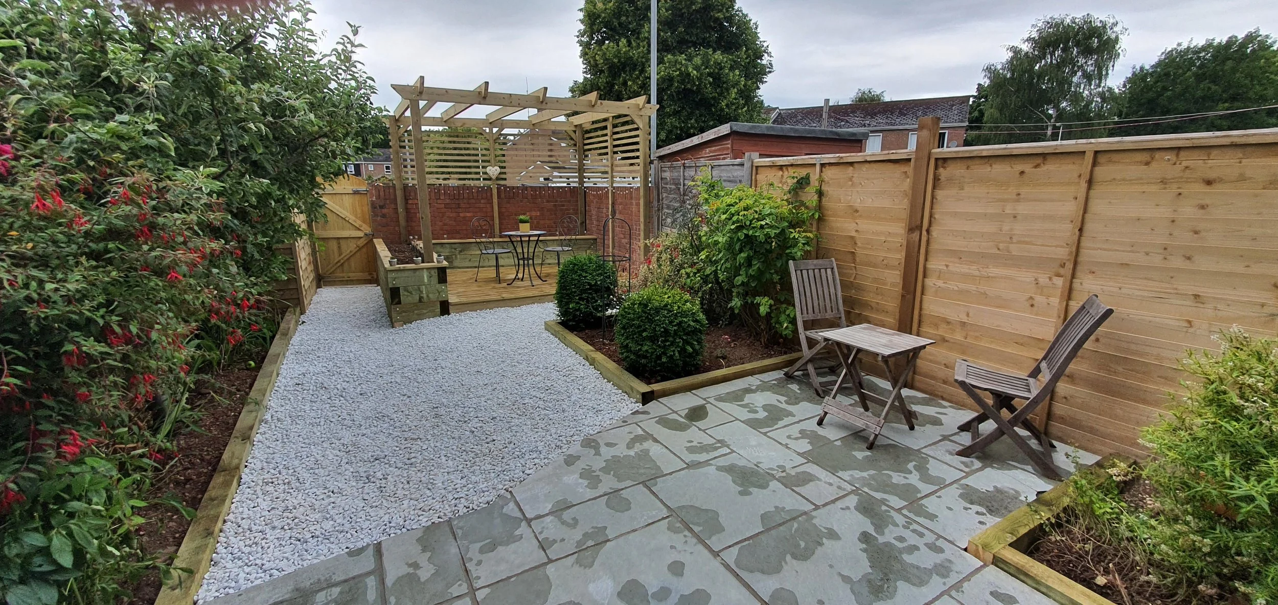 A tidy backyard with a gravel pathway, a wooden fence, and a small patio area with wooden chairs and a table. There is a raised garden bed with green shrubs and a wooden pergola in the background. The yard is bordered by flowering plants and trees.