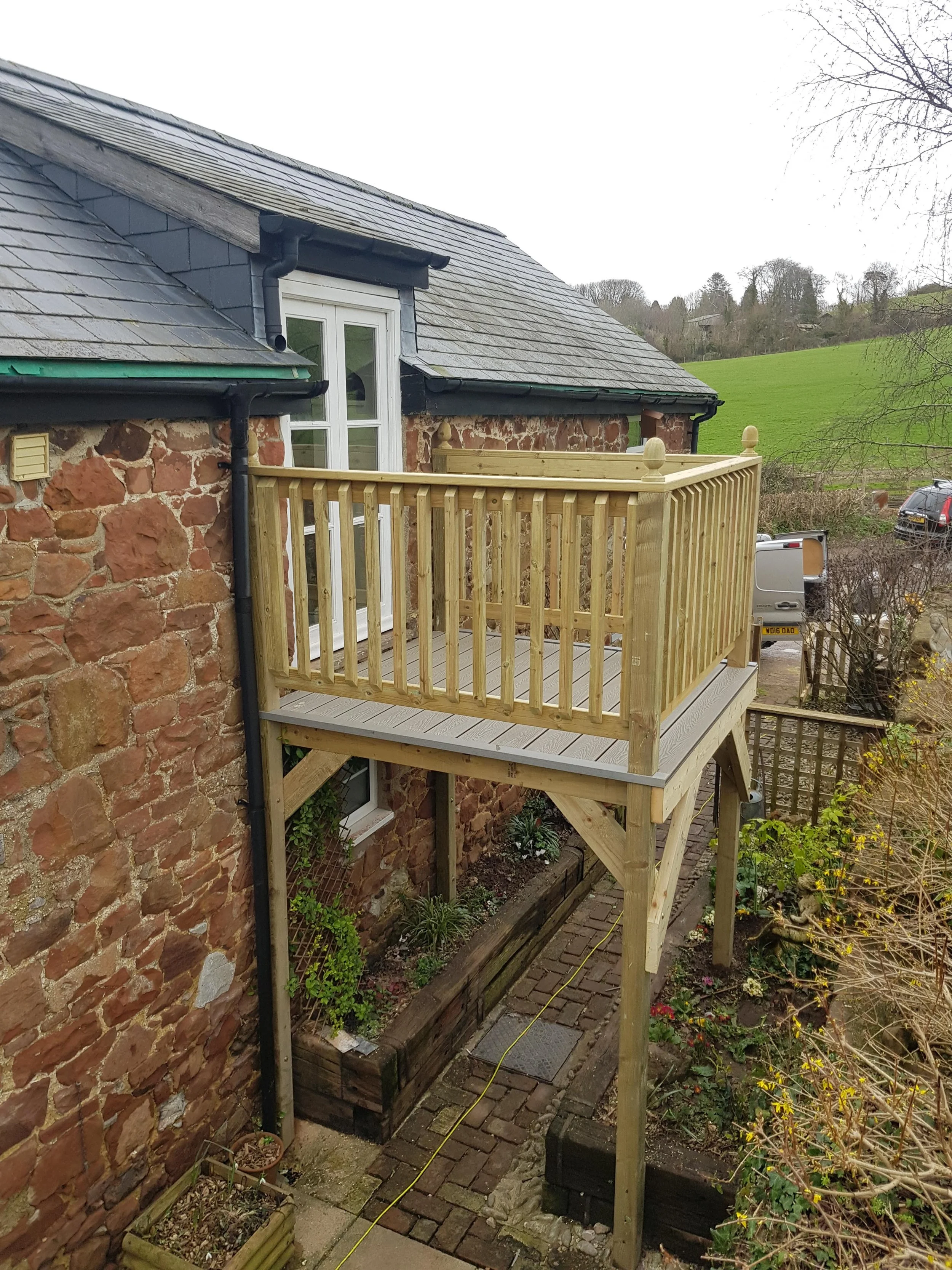New wooden balcony attached to a brick house, overlooking a garden and a parking lot with cars and a green hill in the background.