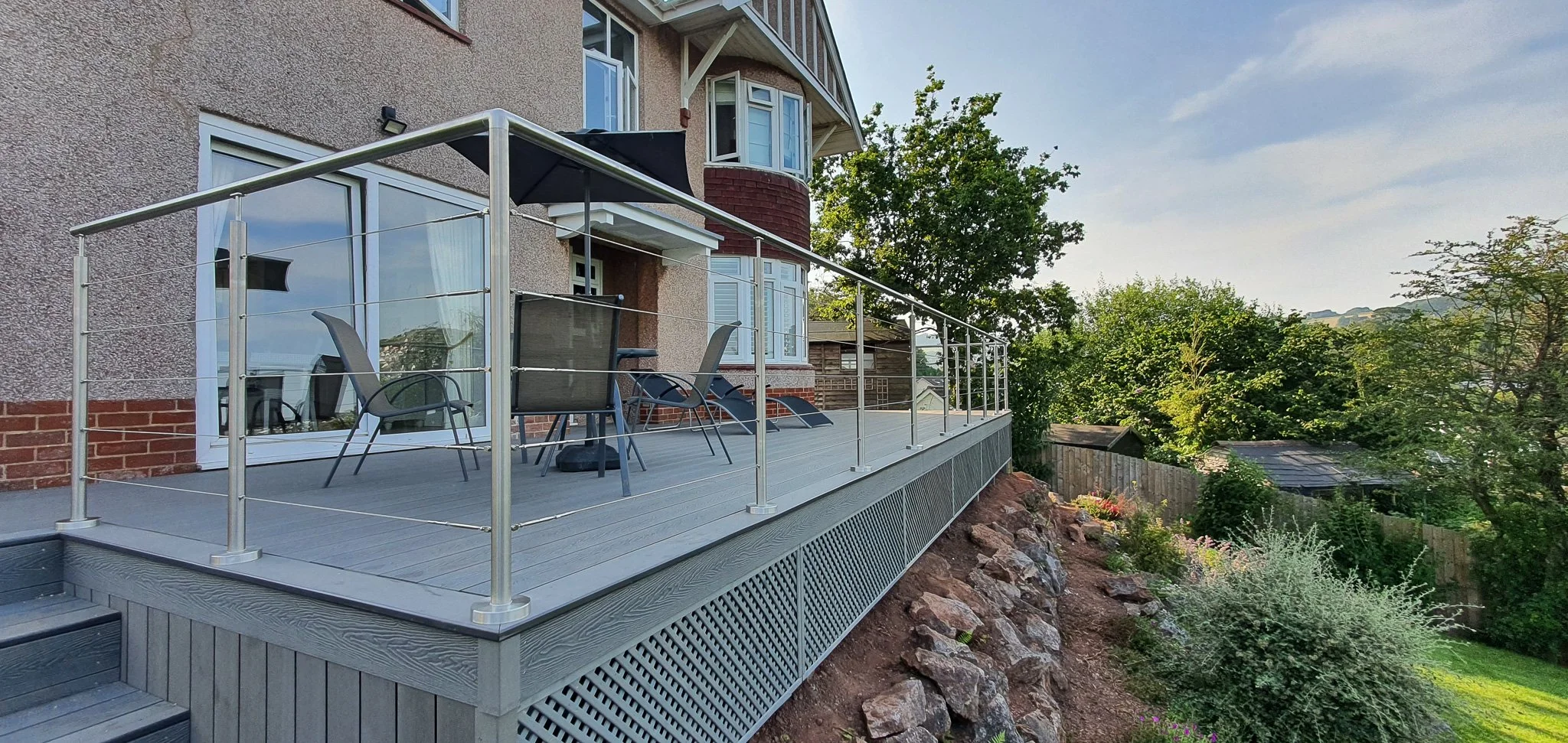 A spacious backyard deck attached to a house, equipped with outdoor chairs, a table, and a sun umbrella, overlooking a lush garden with trees and a wooden fence, under a partly cloudy sky during daylight.