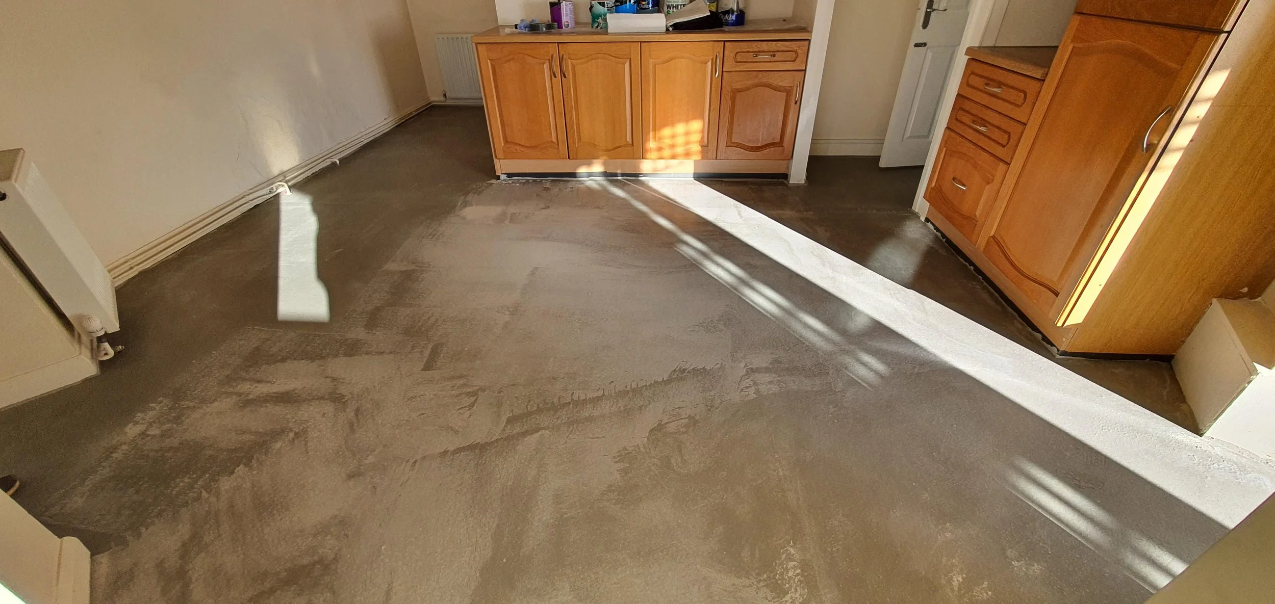 Empty room with a kitchen area, new flooring being installed, sunlight casting shadows on the floor, wooden cabinets, and shelves.