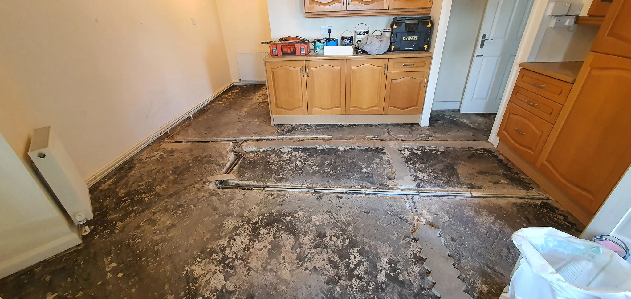 Kitchen floor removed, revealing subfloor and plumbing pipes, with wooden cabinets and some tools and items on the counter.