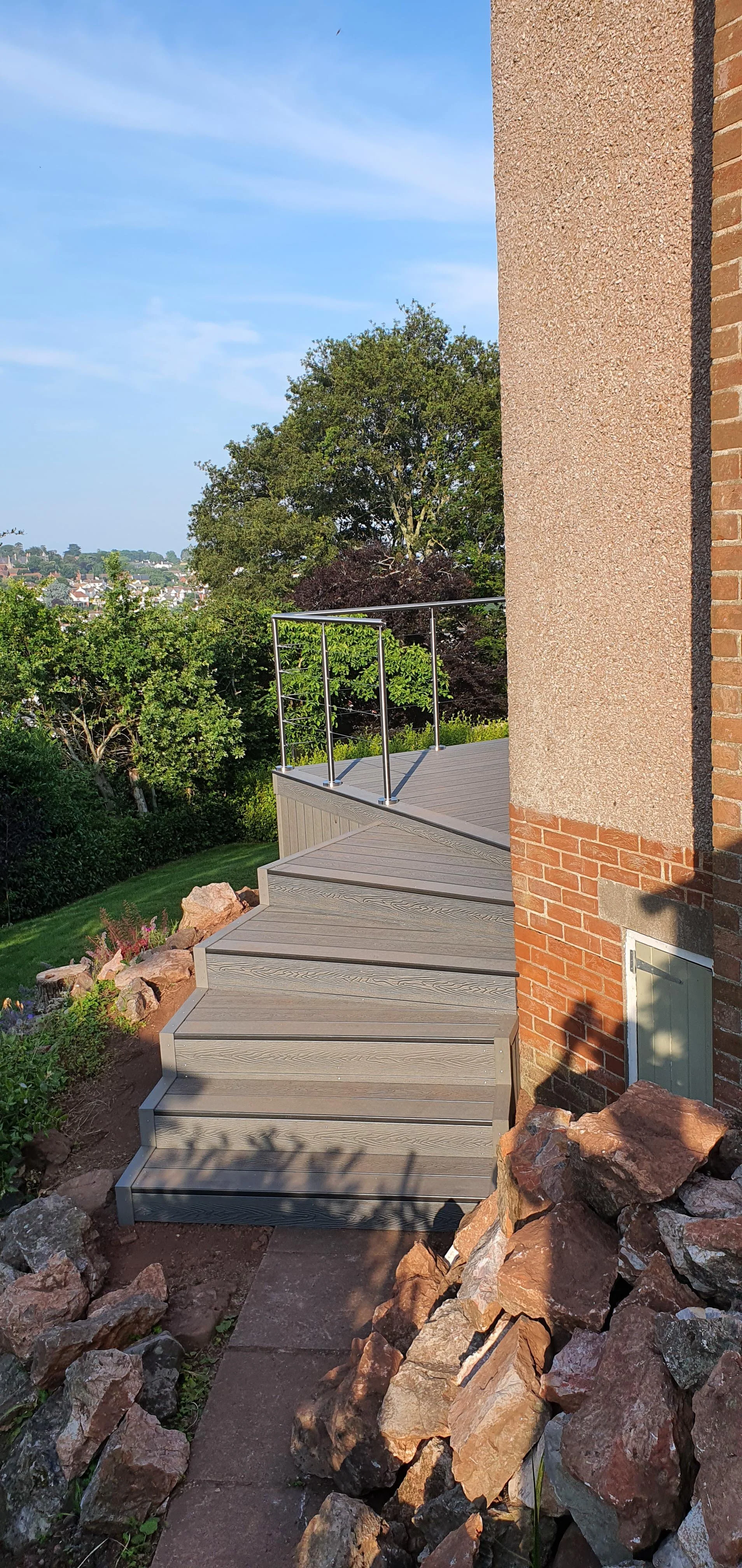 Exterior view of a house with a stairs leading to a deck, a grassy yard with shrubs, and trees in the background under a blue sky.