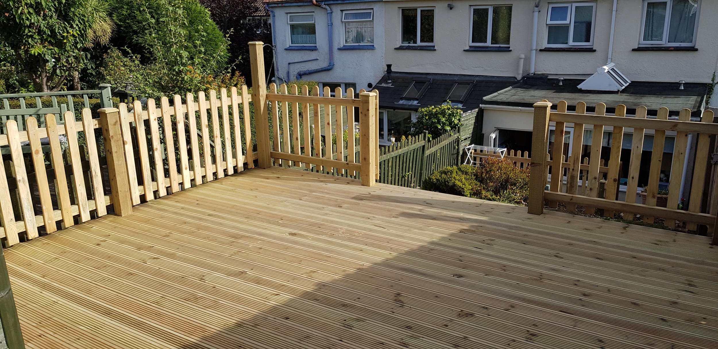 Newly built wooden deck with a railing, surrounded by a garden and neighboring houses.