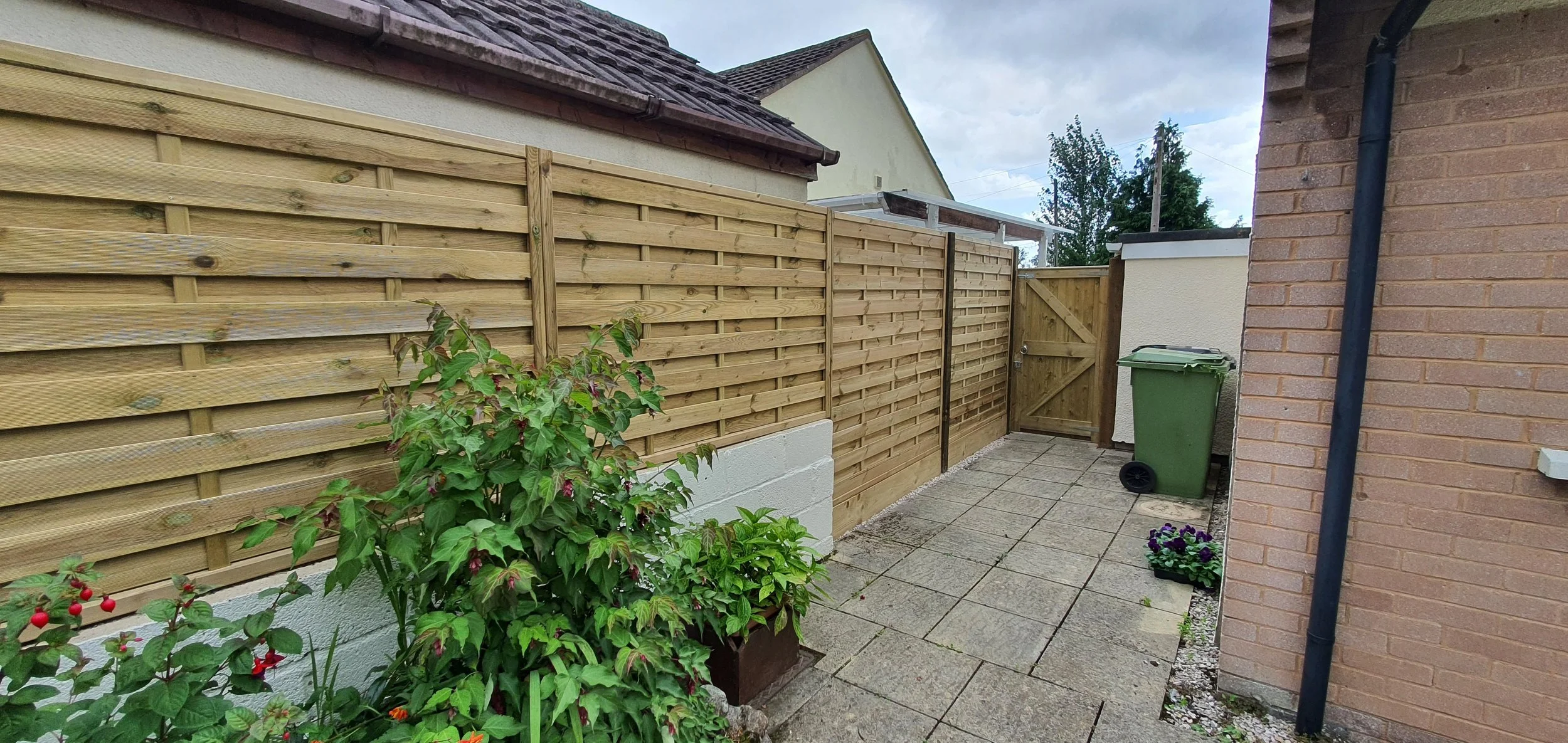 Small backyard with a paved patio, wooden fence, a green trash bin, and potted plants near the wall of a brick house.