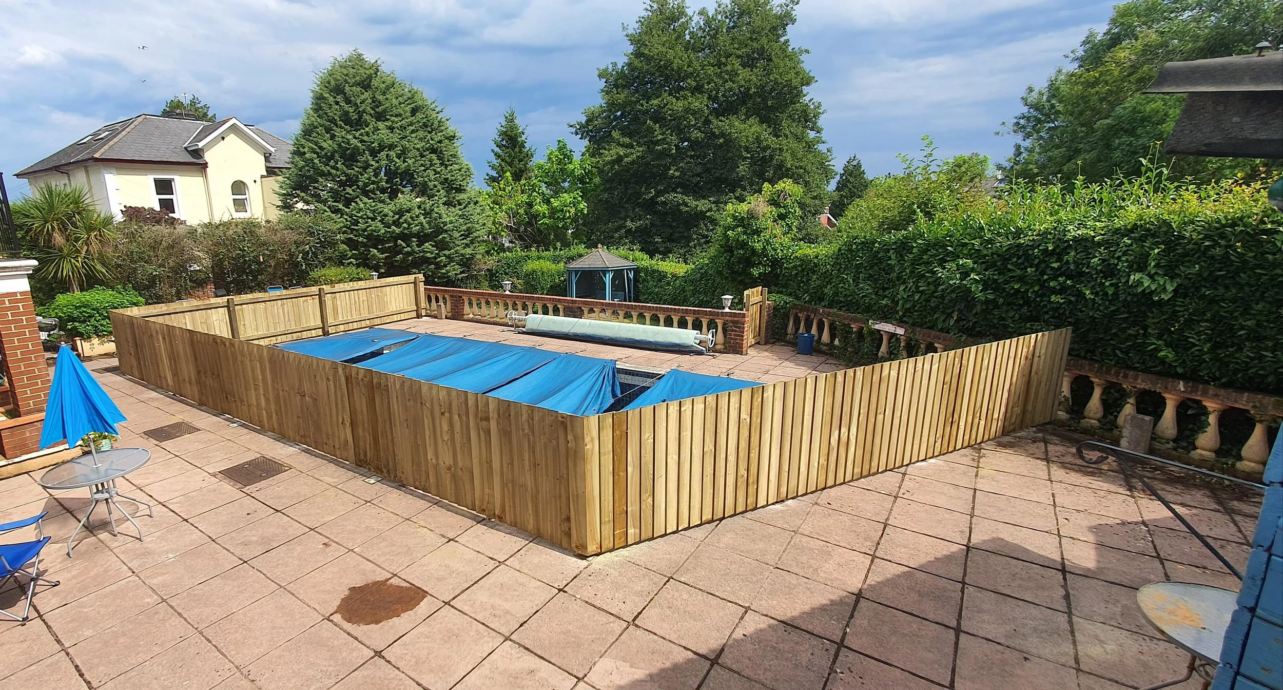 Backyard patio with a fenced-in swimming pool covered with a blue tarp, surrounded by paving stones, patio furniture, umbrellas, and lush green trees.