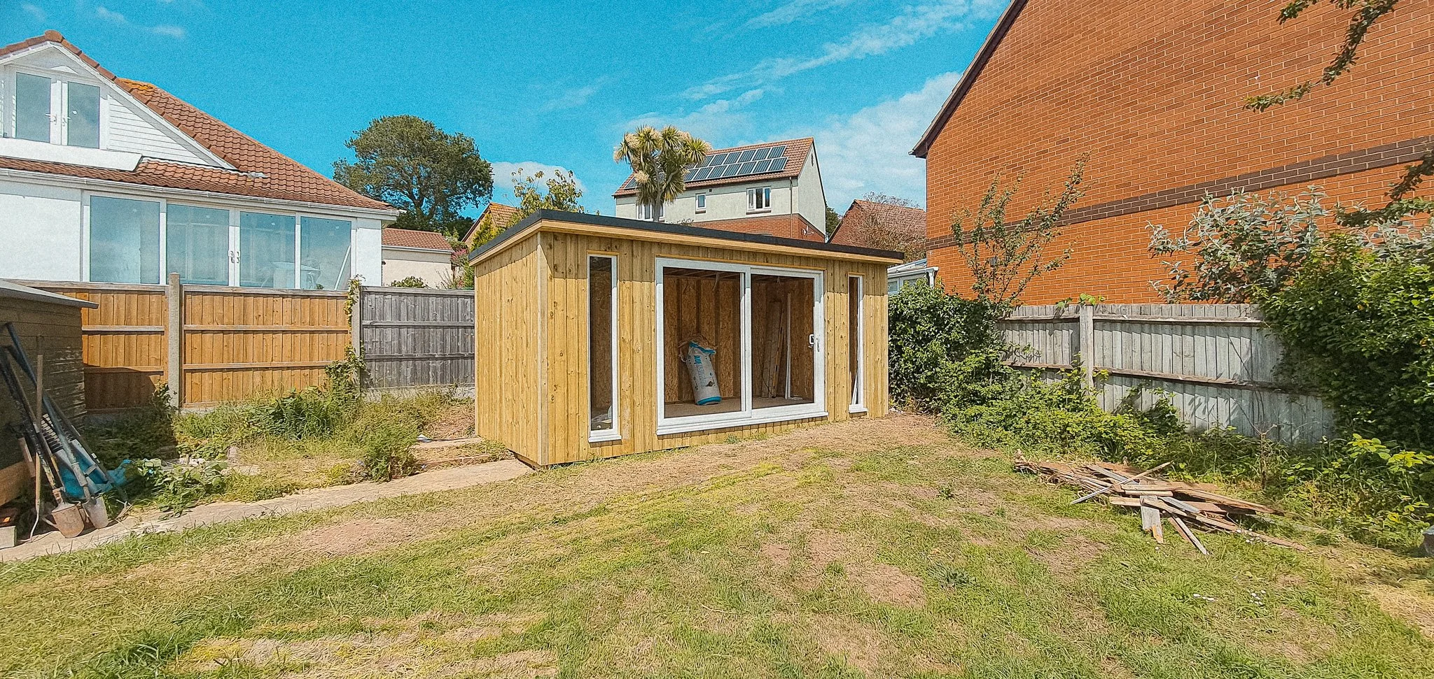A small wooden shed under construction in a backyard, with a large sliding glass door and side windows, surrounded by a grassy area and neighboring houses.