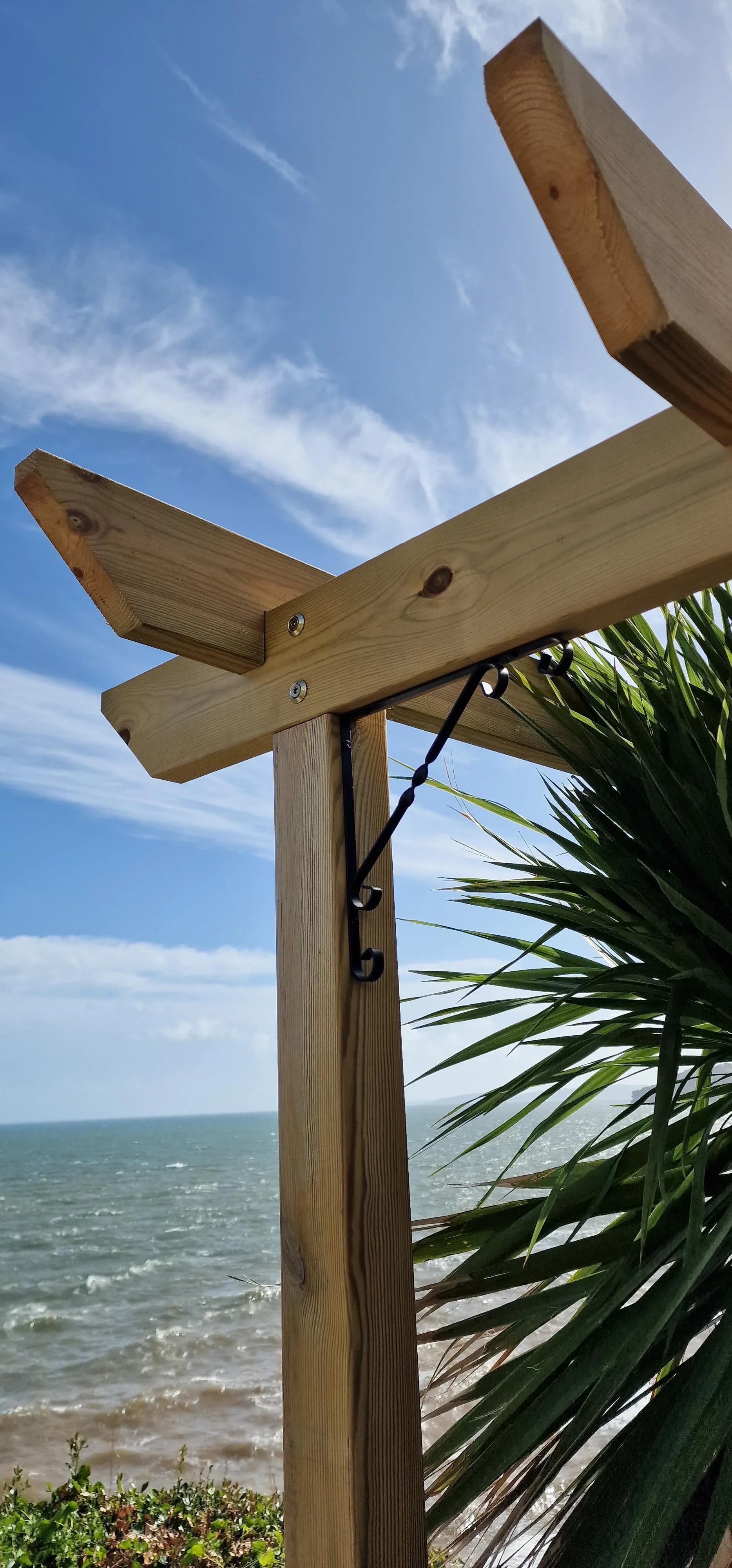 A wooden structure with diagonal pieces, metal brackets, and hooks near a beach, with ocean waves, blue sky, and clouds in the background, and green foliage.