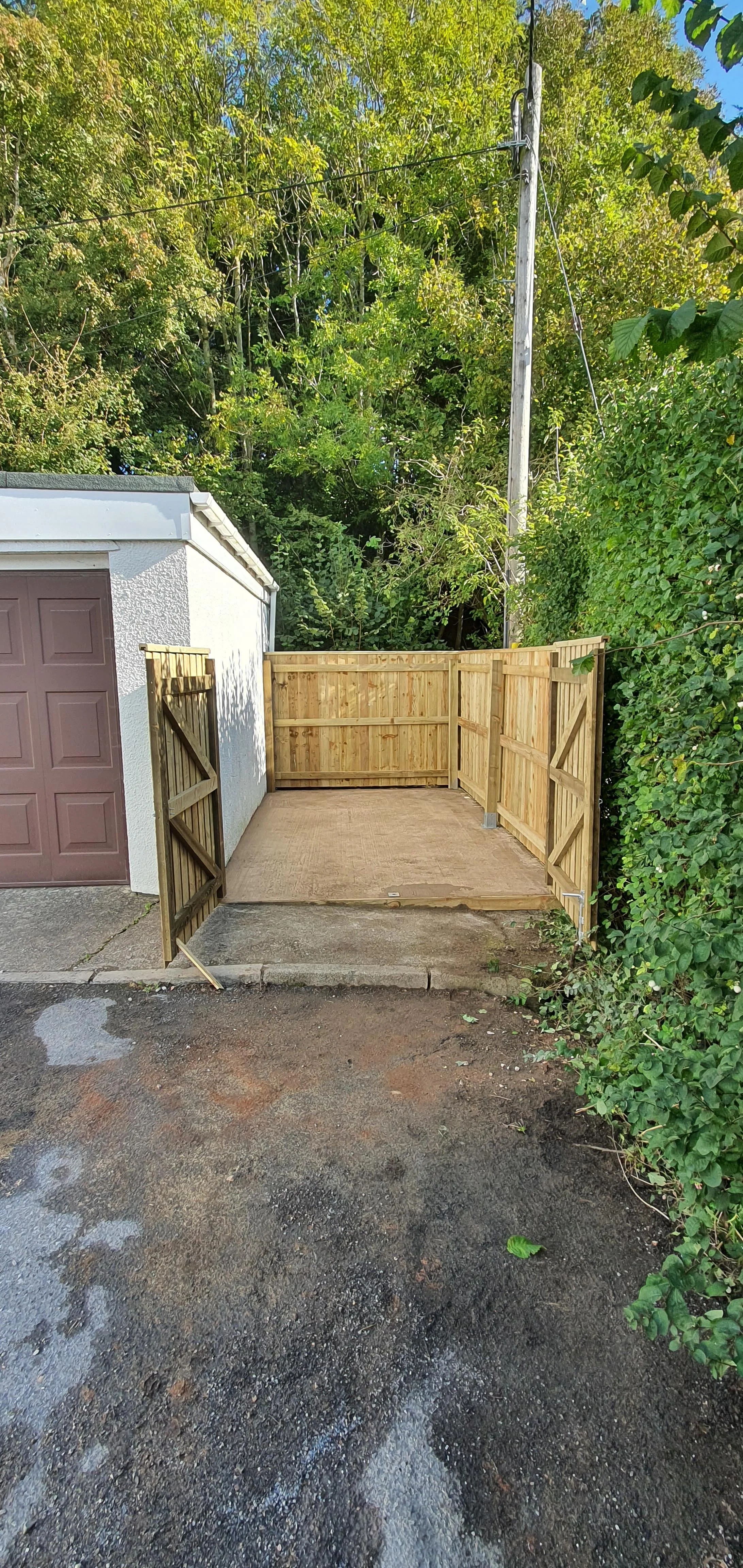 A newly built wooden fence surrounds a small concrete patio area next to a garage, with green trees and bushes in the background.