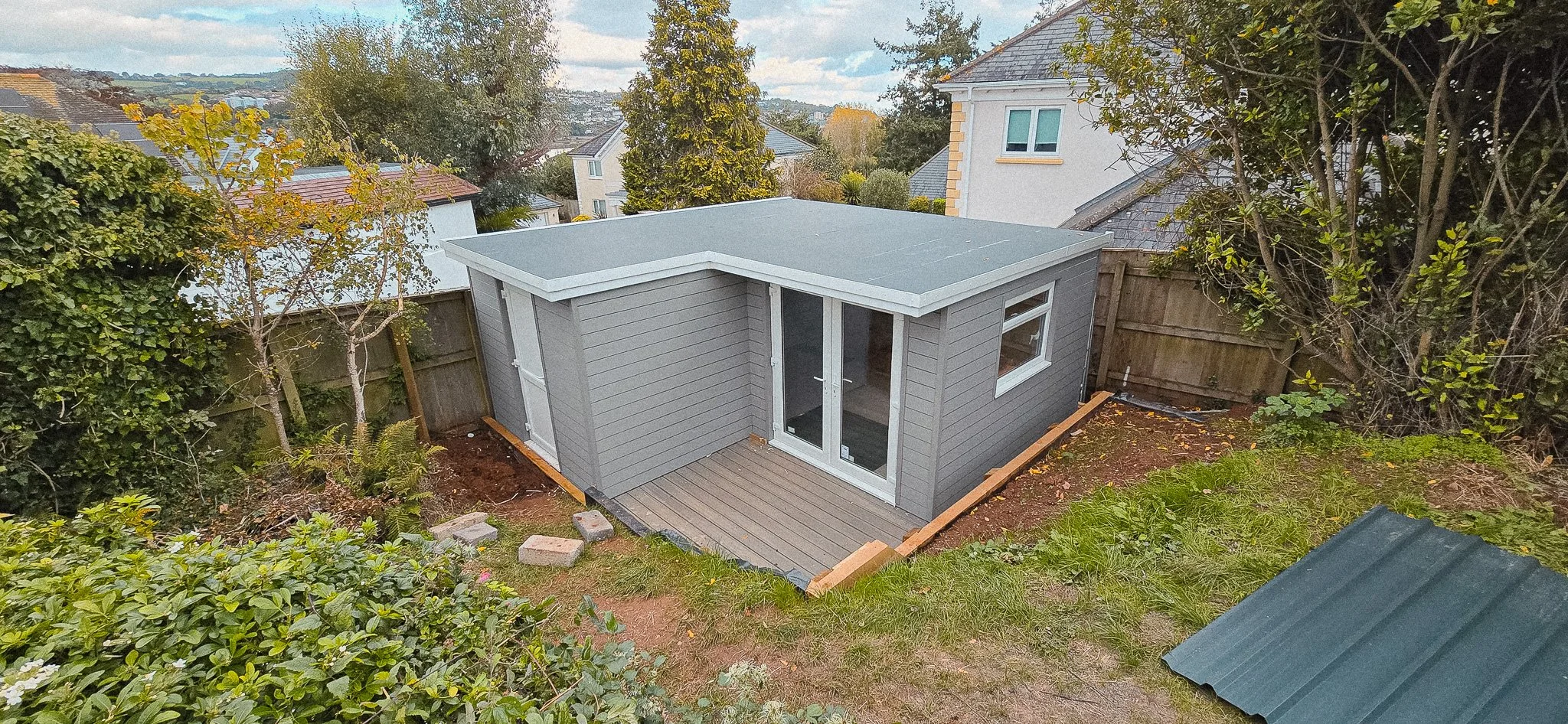 A small grey backyard shed with a flat roof, located in a fenced backyard with trees and bushes, and a grassy area in front and to the side.