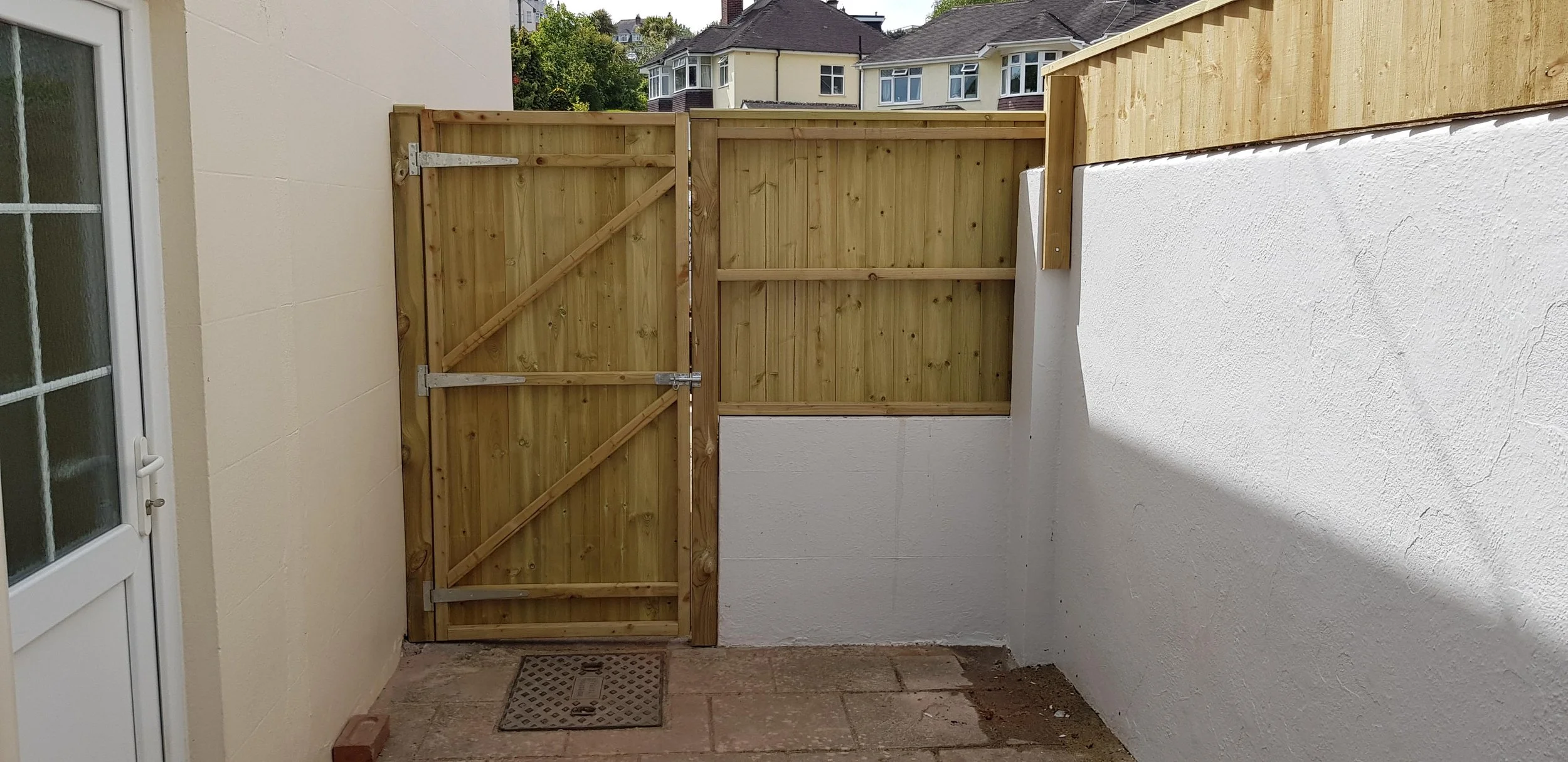 A small outdoor patio area with a new wooden gate and fencing, white and cream walls, a brick and tile ground, and neighboring houses visible in the background.
