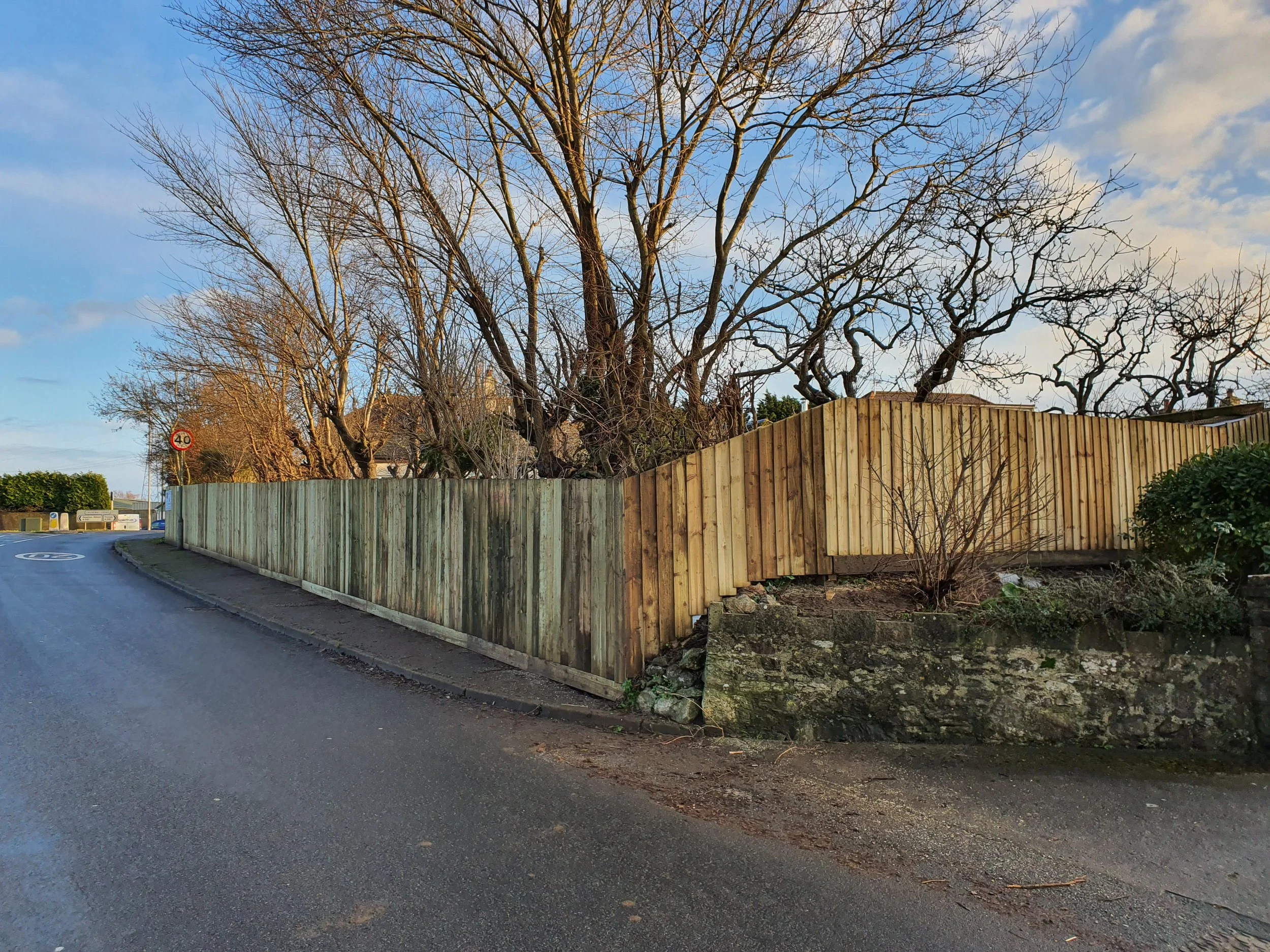 A curved asphalt road with a yellow wooden fence on the right side, behind which leafless trees and some bushes are visible. The sky is partly cloudy with patches of blue.