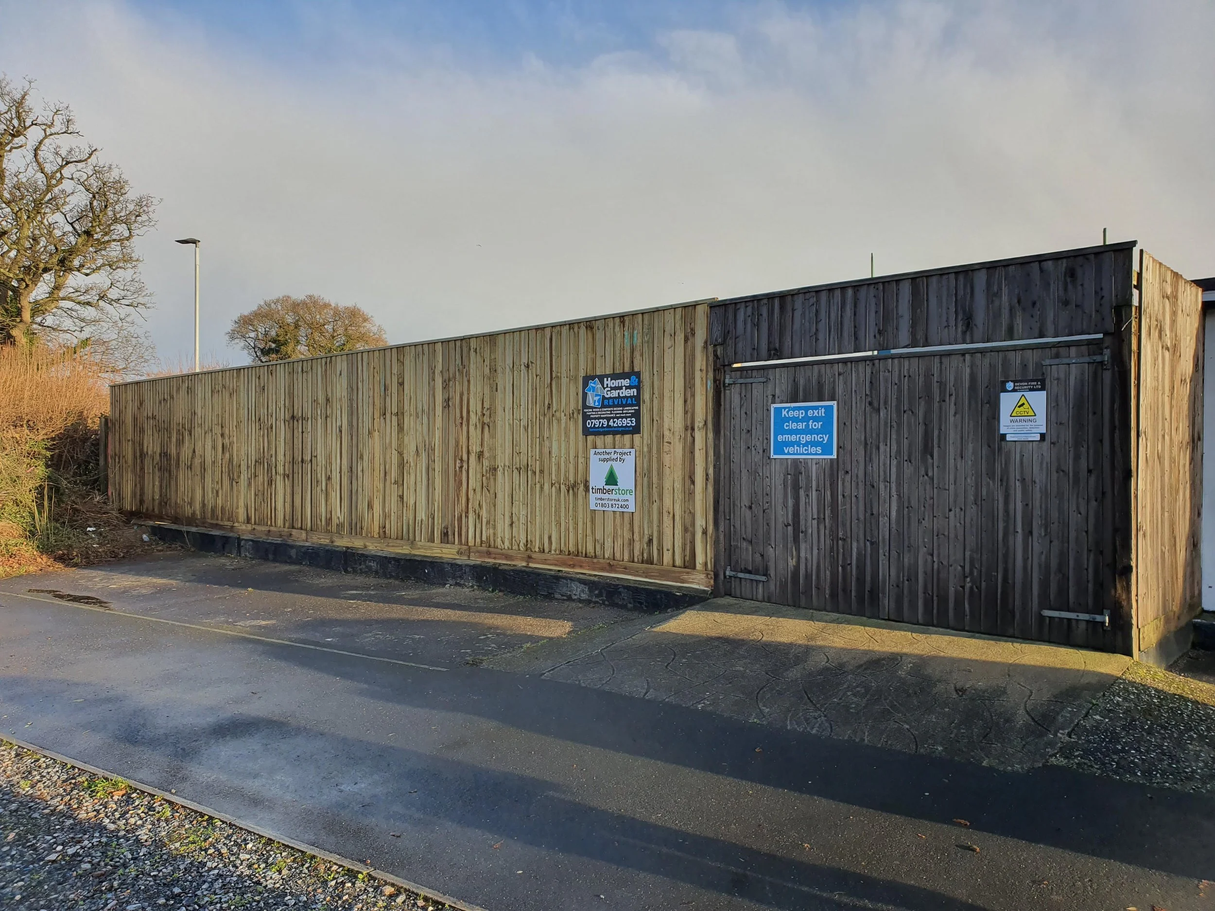 Wooden fence and gate with signs, next to a paved road and trees in the background.