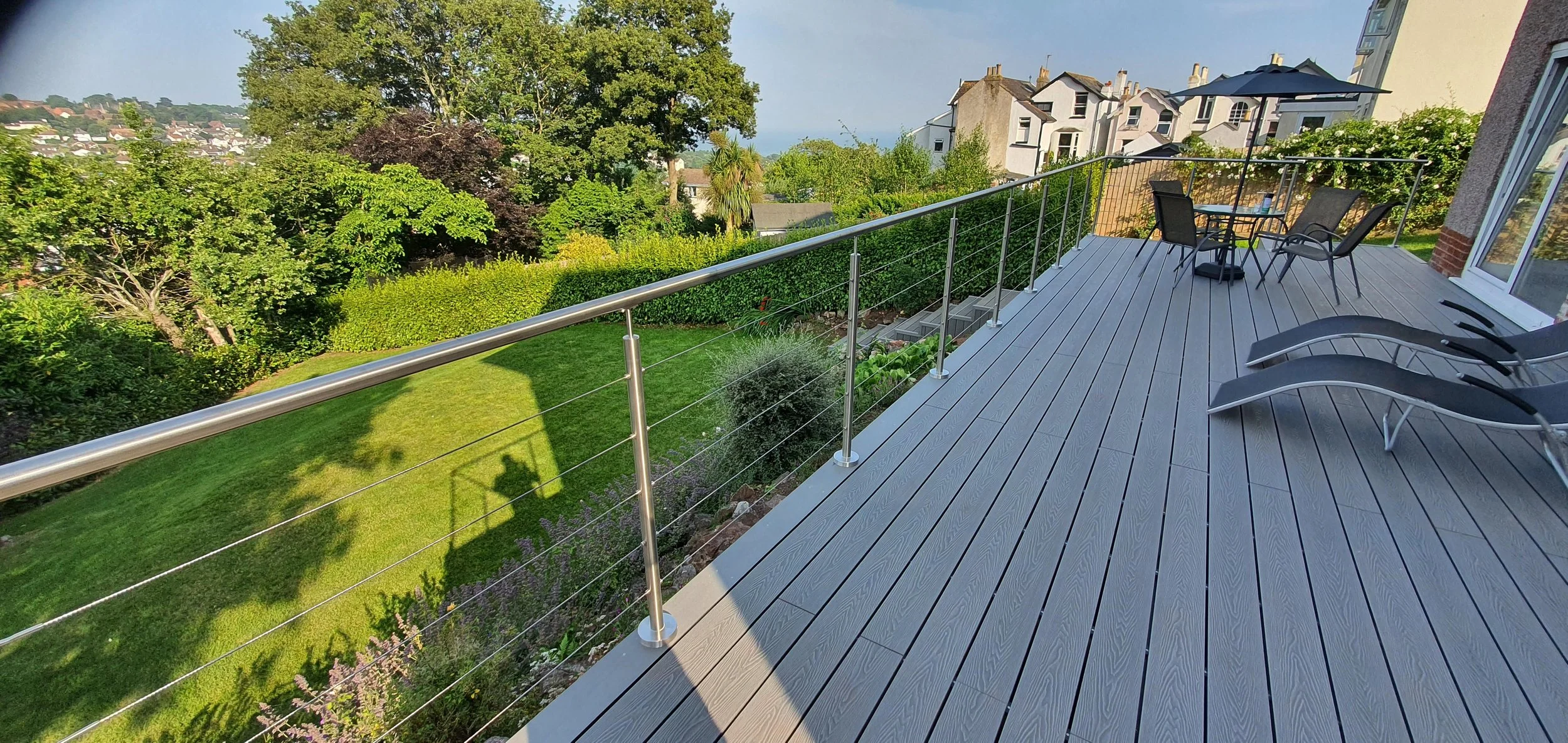 A wooden deck with patio furniture and lounge chairs overlooking a lush garden with trees and houses in the background.