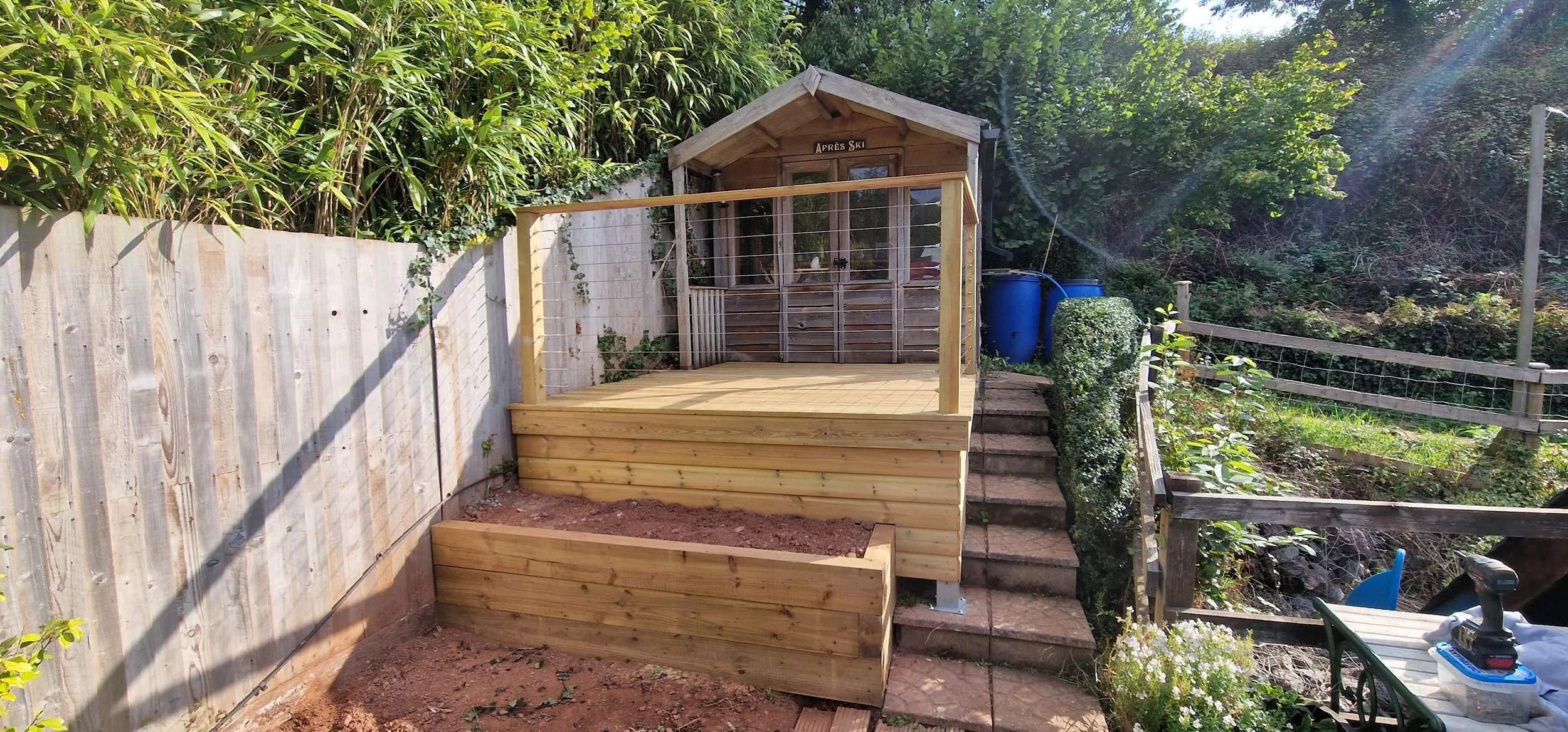 A small wooden deck with a safety rail, built in a backyard with stair access, leading to a small wooden shed labeled 'Après Ski'. There are blue rain barrels next to the shed, and a garden with trees and plants surrounds the area.