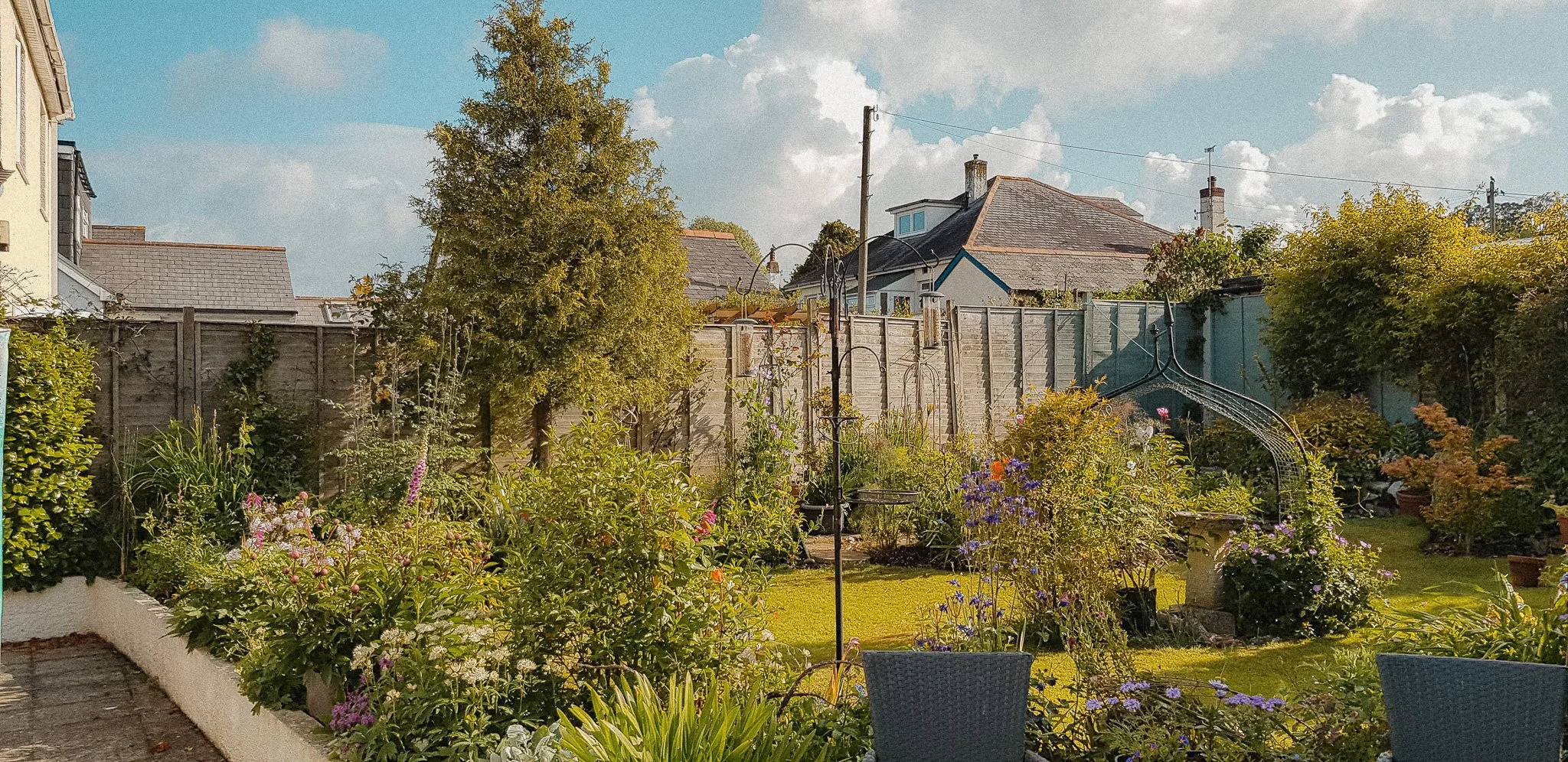 A lush garden with blooming flowers, green shrubs, a small lawn, and an outdoor swing set, enclosed by a wooden fence under a partly cloudy sky.