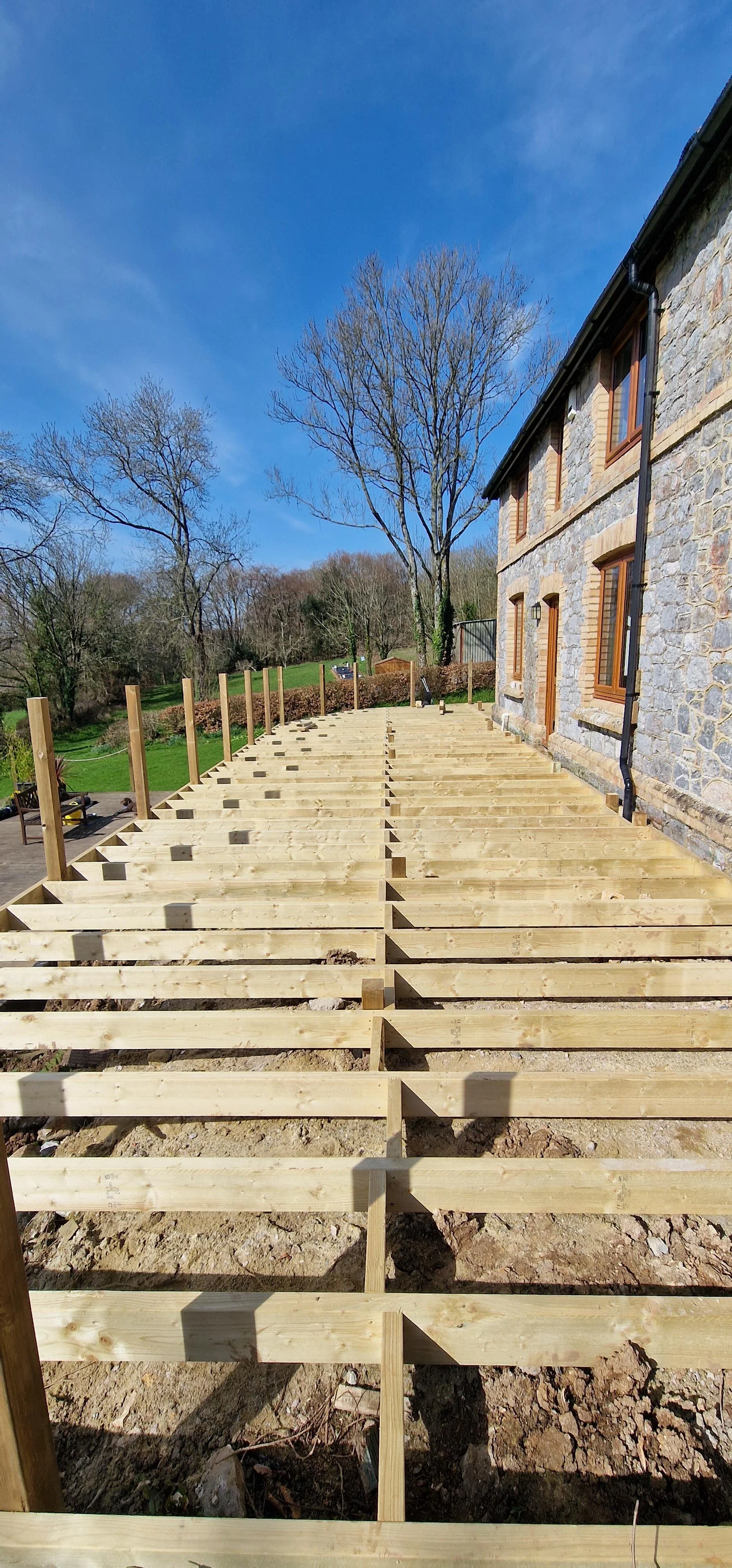 Wooden deck framing construction in progress beside a stone house, with leafless trees and a blue sky in the background.