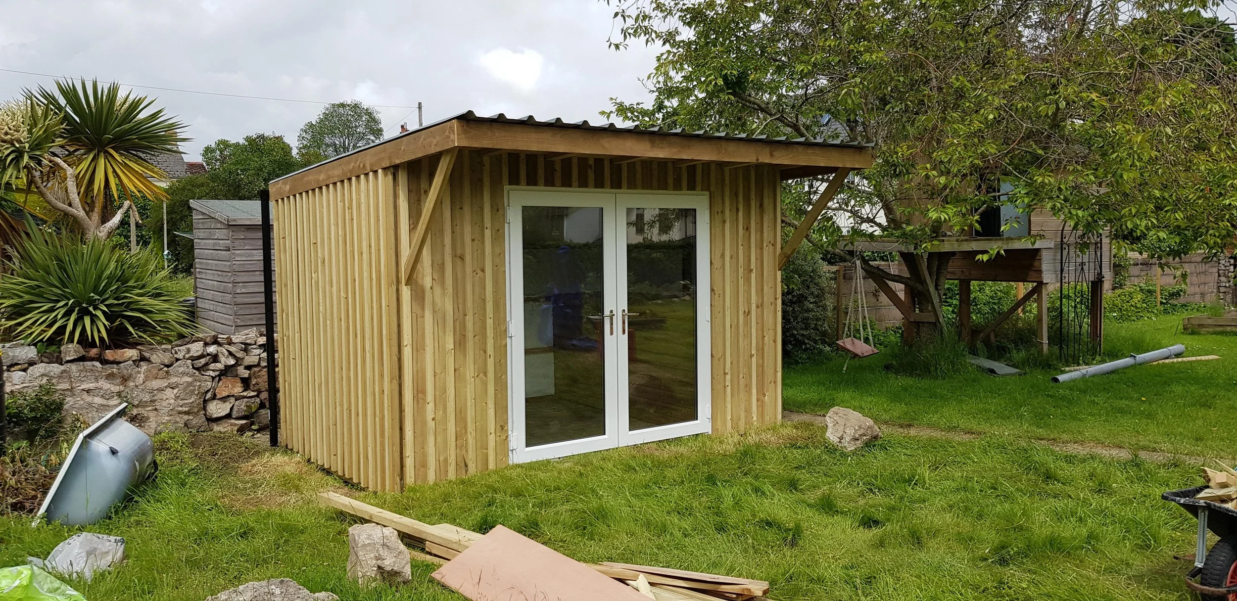 Small wooden shed with glass double doors, situated in a grassy backyard with trees and plants, construction materials and tools scattered around.