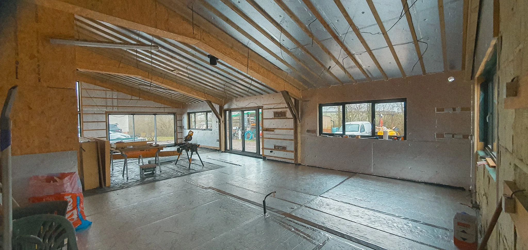 Interior of a house under construction, showing exposed wooden framing, partially finished walls, large windows, and a vaulted ceiling. Construction tools and supplies are scattered around.
