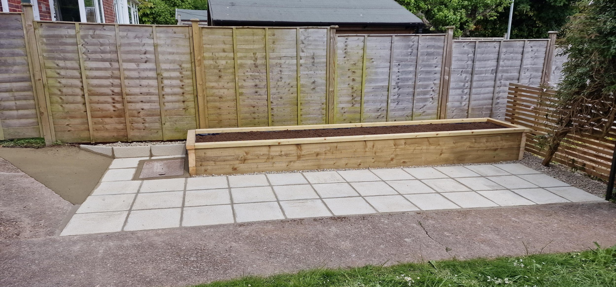 A newly built wooden garden bed on a patio, surrounded by a wooden fence and paved stone tiles.