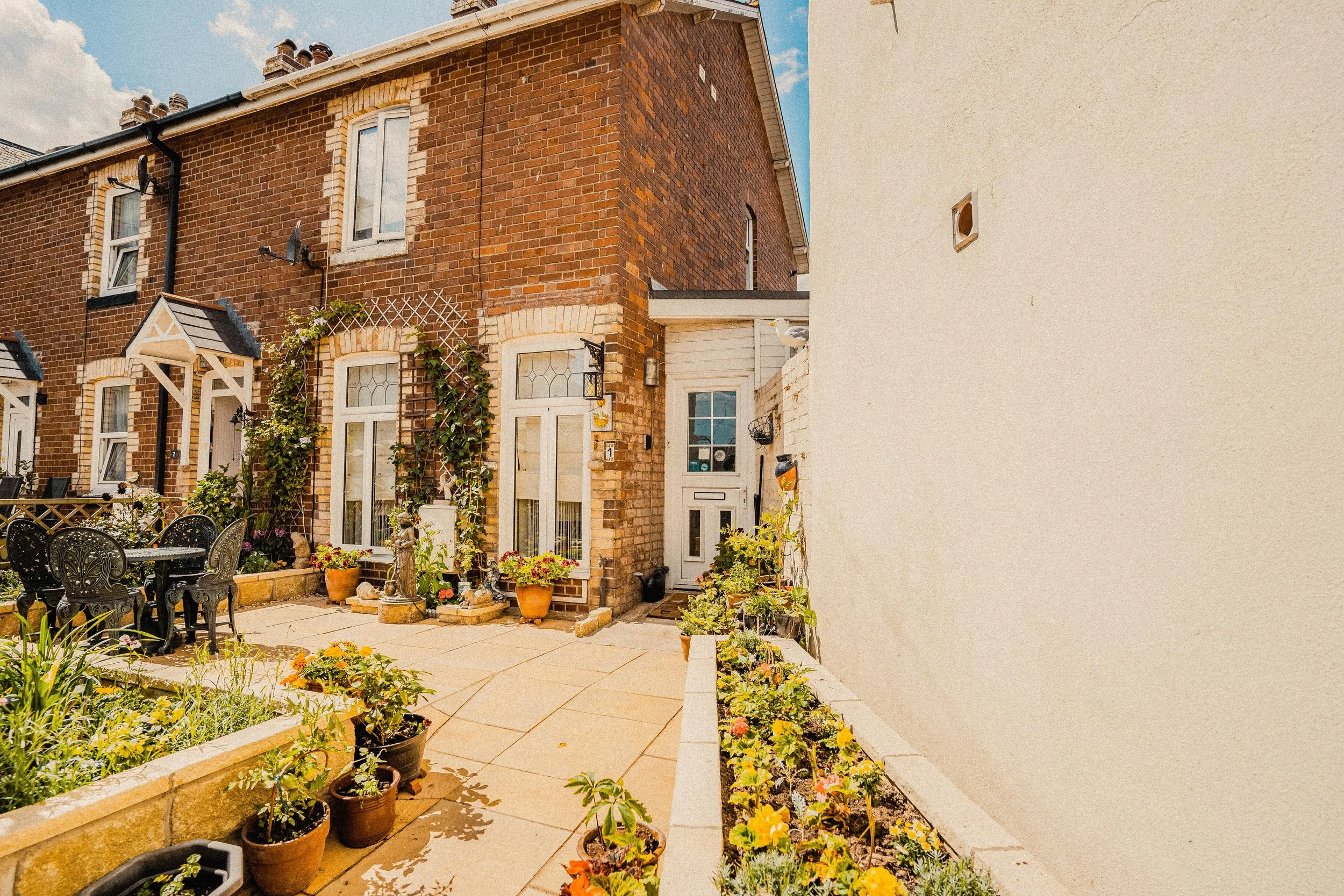 Backyard garden with potted plants, flowers, and outdoor seating, adjacent to a brick house with a white door and windows, under a partly cloudy sky.