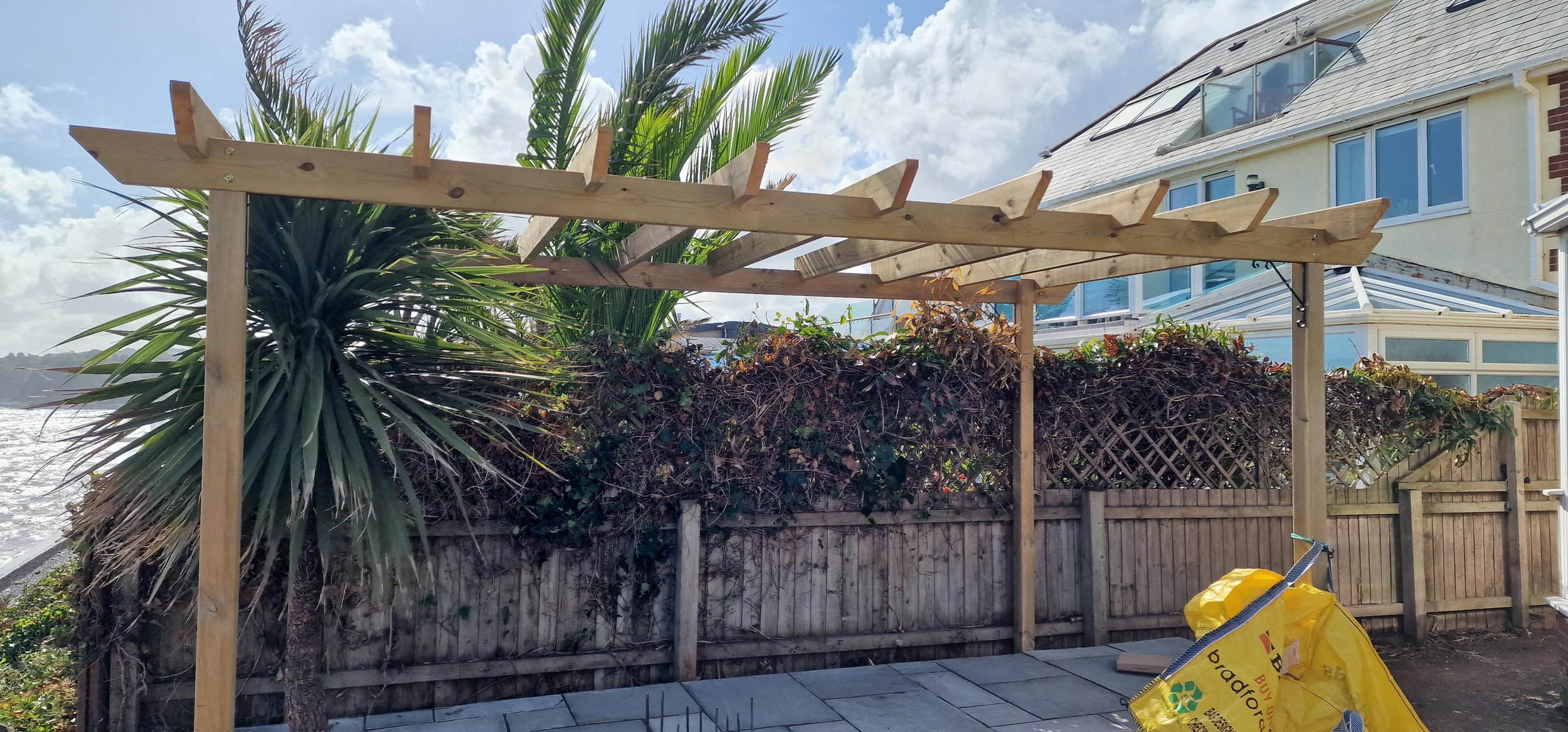 A wooden framework for a pergola or roof extension under construction in a backyard, with a large palm and ivy-covered fence, a house with large windows, and construction materials on the ground.