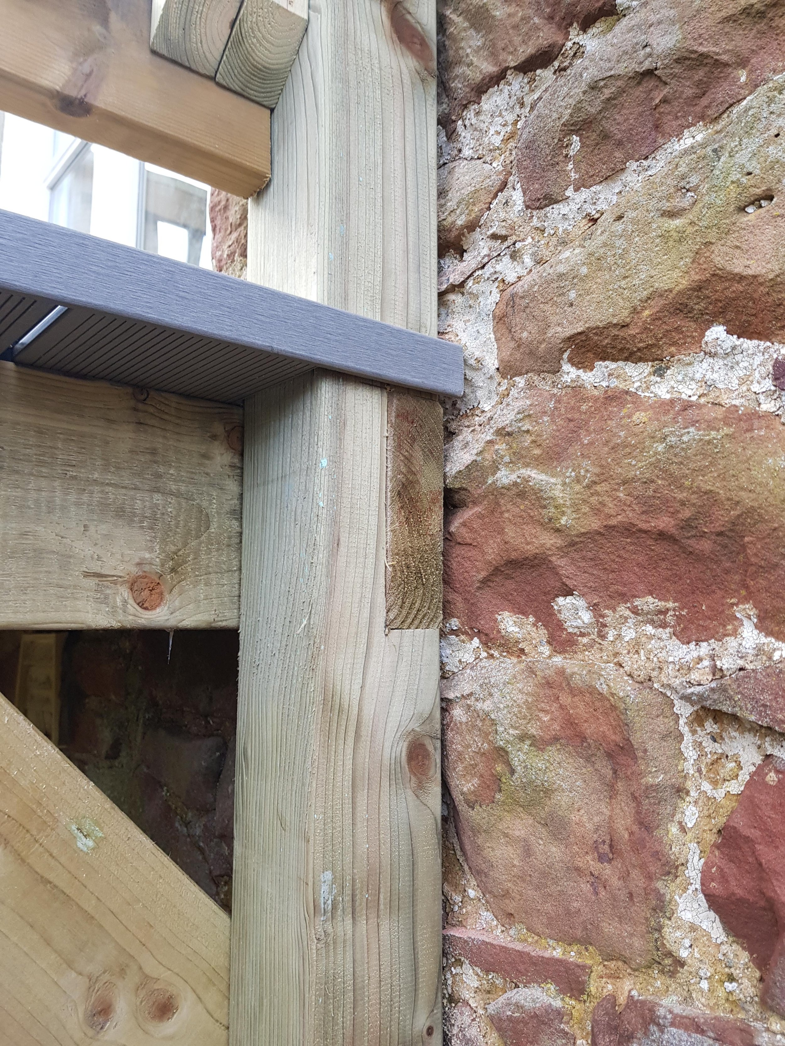 Close-up of a wooden structure with a metal angle bracket against a red brick wall.