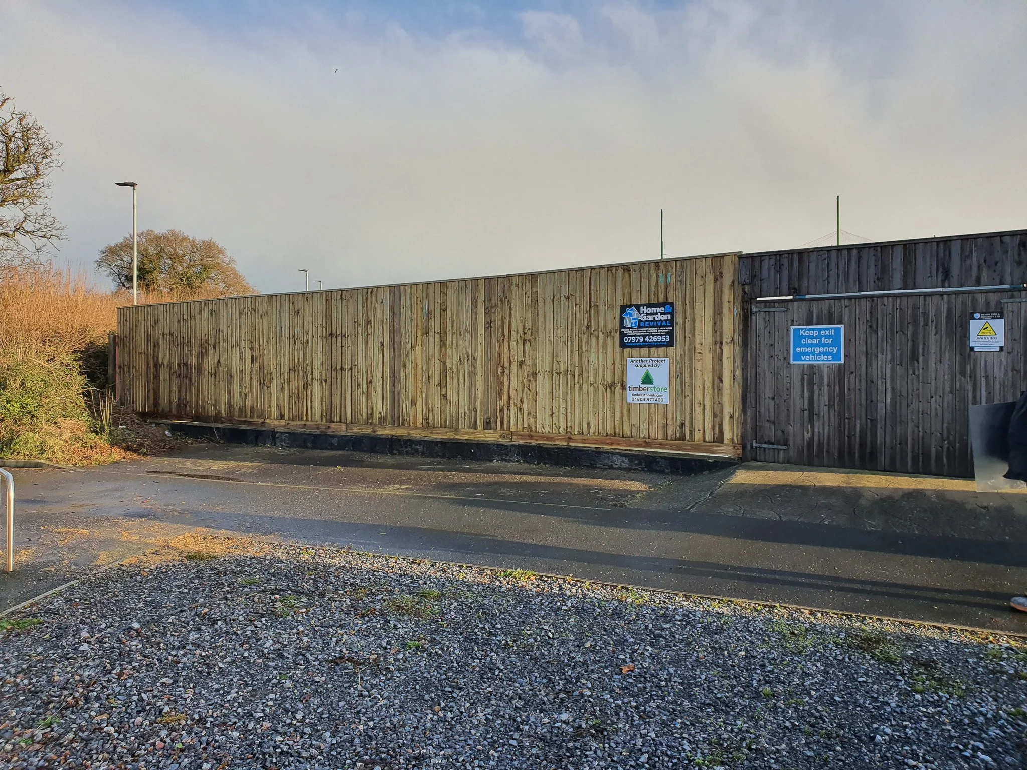Wooden fence with multiple signs, including one for Home & Garden Revival, a keep exit clear sign, and a warning sign, with a cloudy sky and some trees in the background.