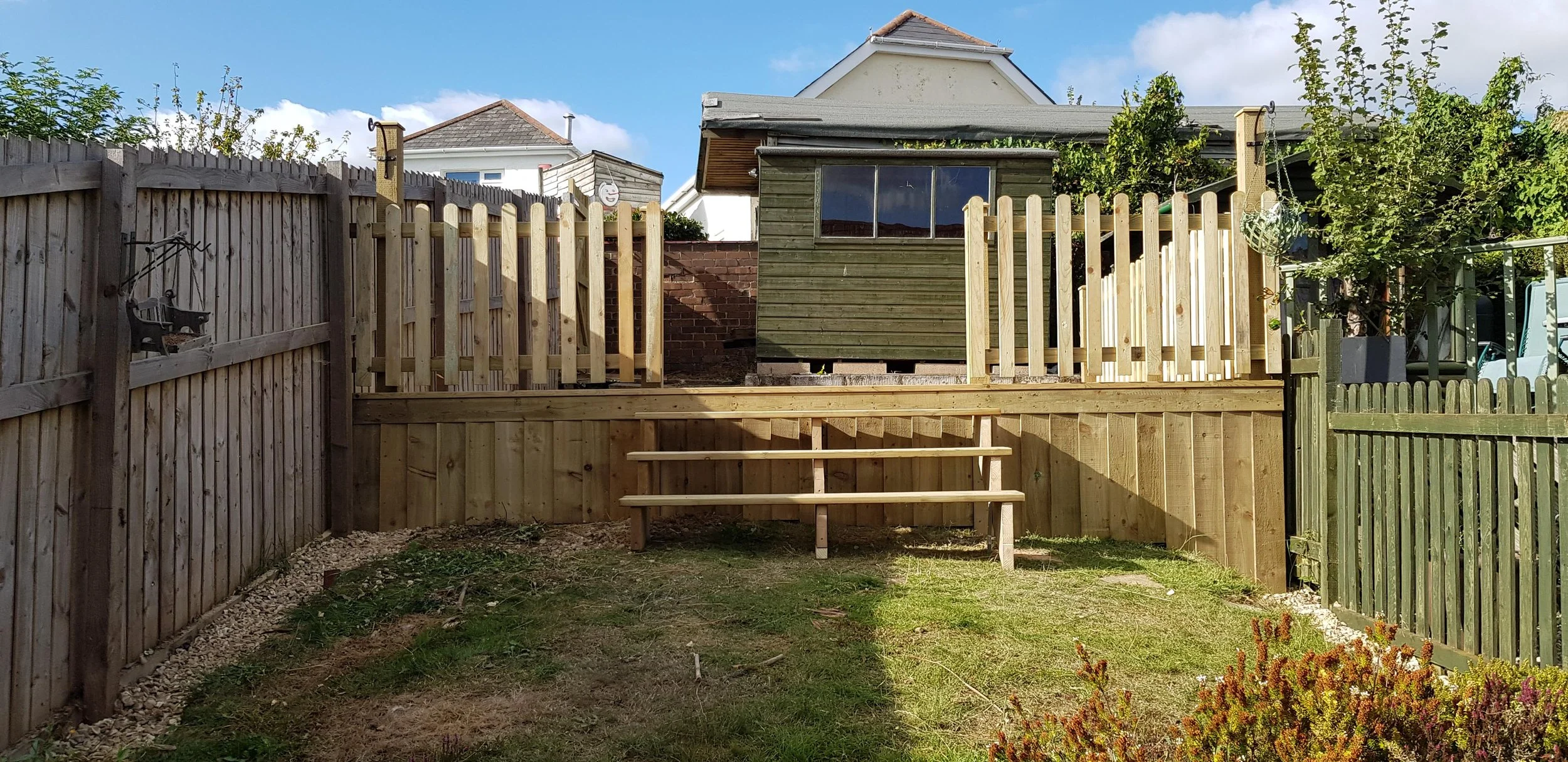 Backyard with wooden fence, small grassy area, and a raised wooden deck with steps, behind a house with a green wooden shed, trees, and neighboring houses visible in the background.