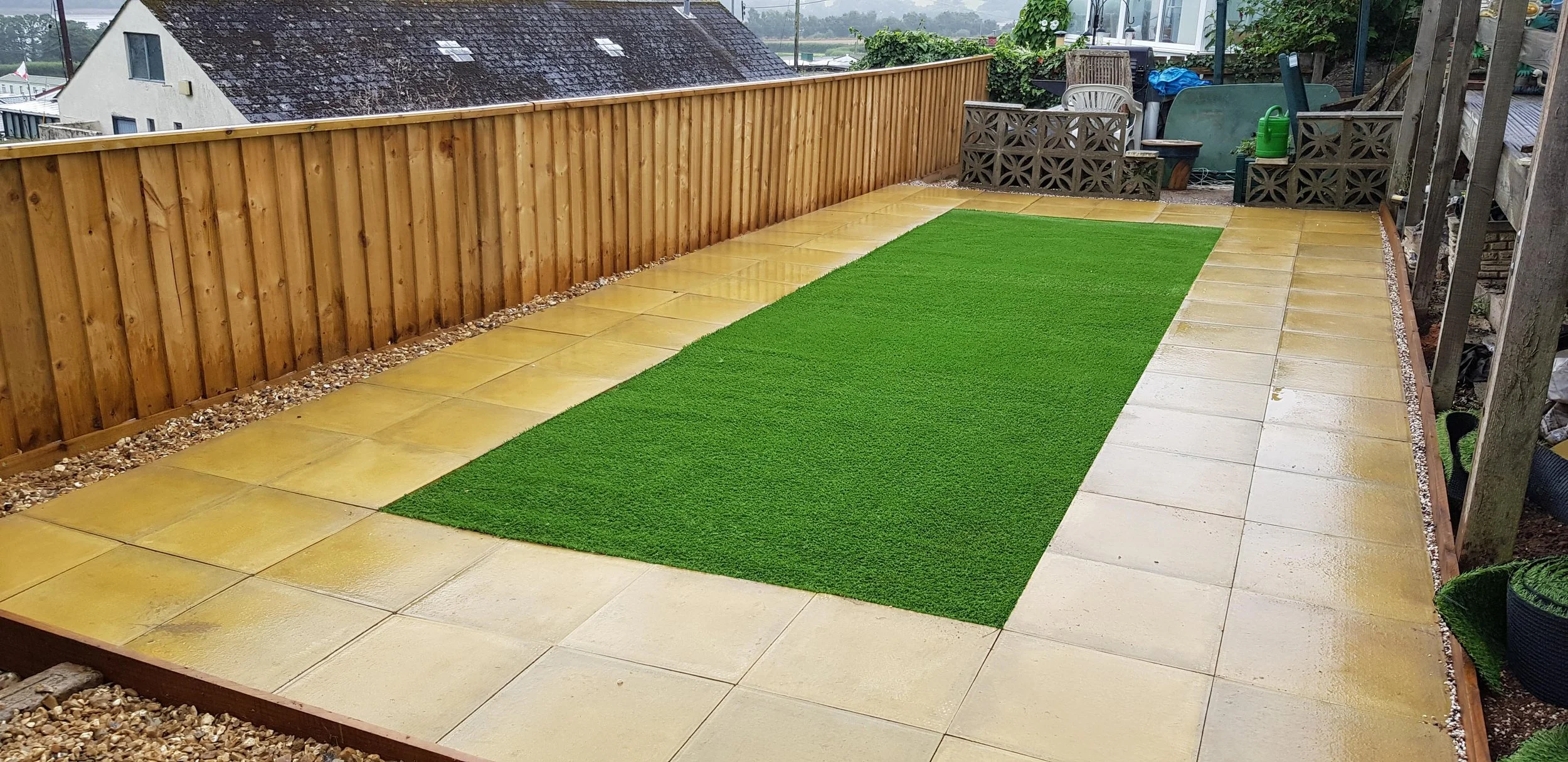 A backyard patio with wet yellow tiles, a green artificial grass patch in the center, a wooden fence on one side, and outdoor furniture with a watering can, plant pots, and other gardening tools in the corner. The area appears to be wet from rain.