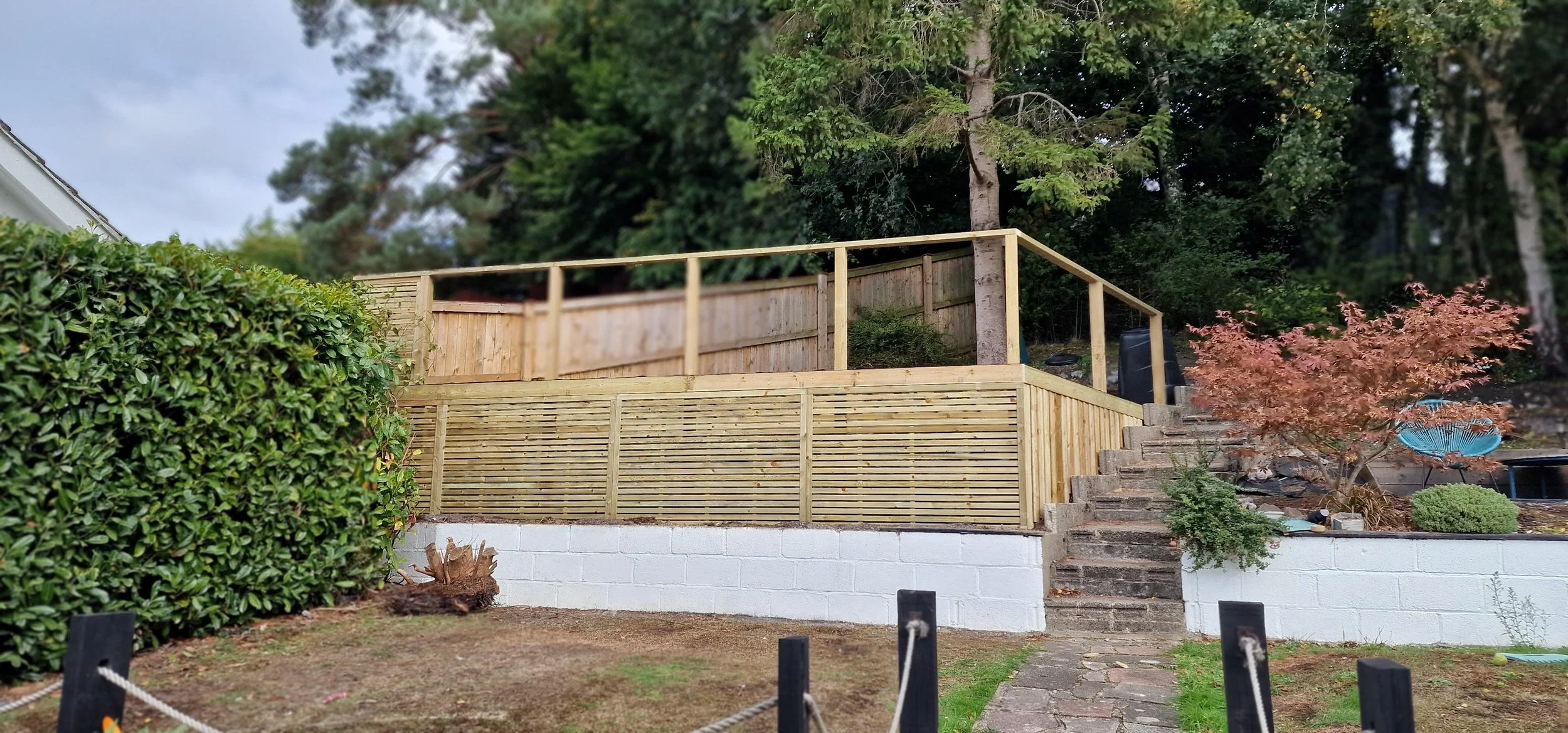 A landscaped backyard with a white painted brick wall, wooden staircase, and wooden fencing, surrounded by trees and bushes.